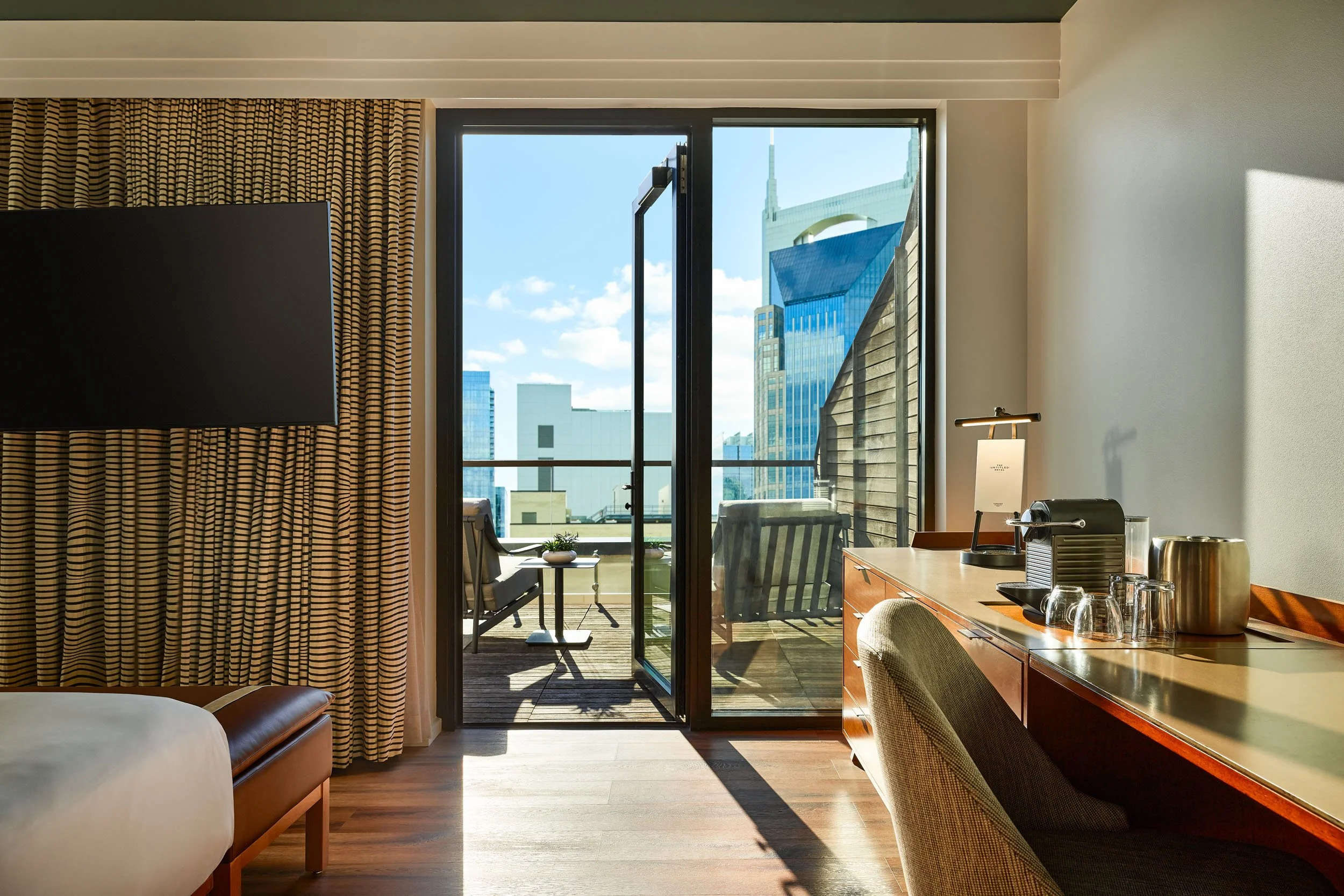 A hotel room with a sliding glass door leading to a balcony overlooking a city skyline with modern high-rise buildings and blue sky. Inside, there's a wooden desk with a lamp, glassware, and a coffee machine, and a curtain with black and beige stripe
