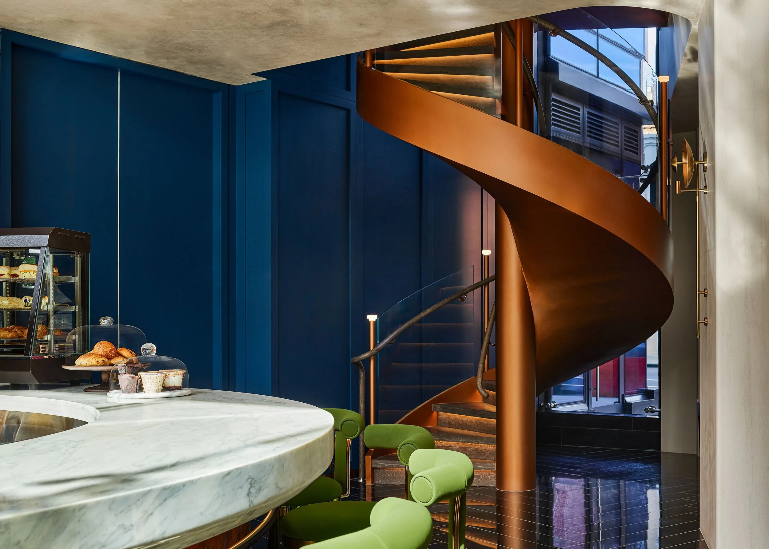 Interior of a modern cafe or restaurant with a marble counter, a glass display case of baked goods, a spiral staircase with a copper finish, and vibrant green chairs around a small table, featuring contemporary design and warm lighting.