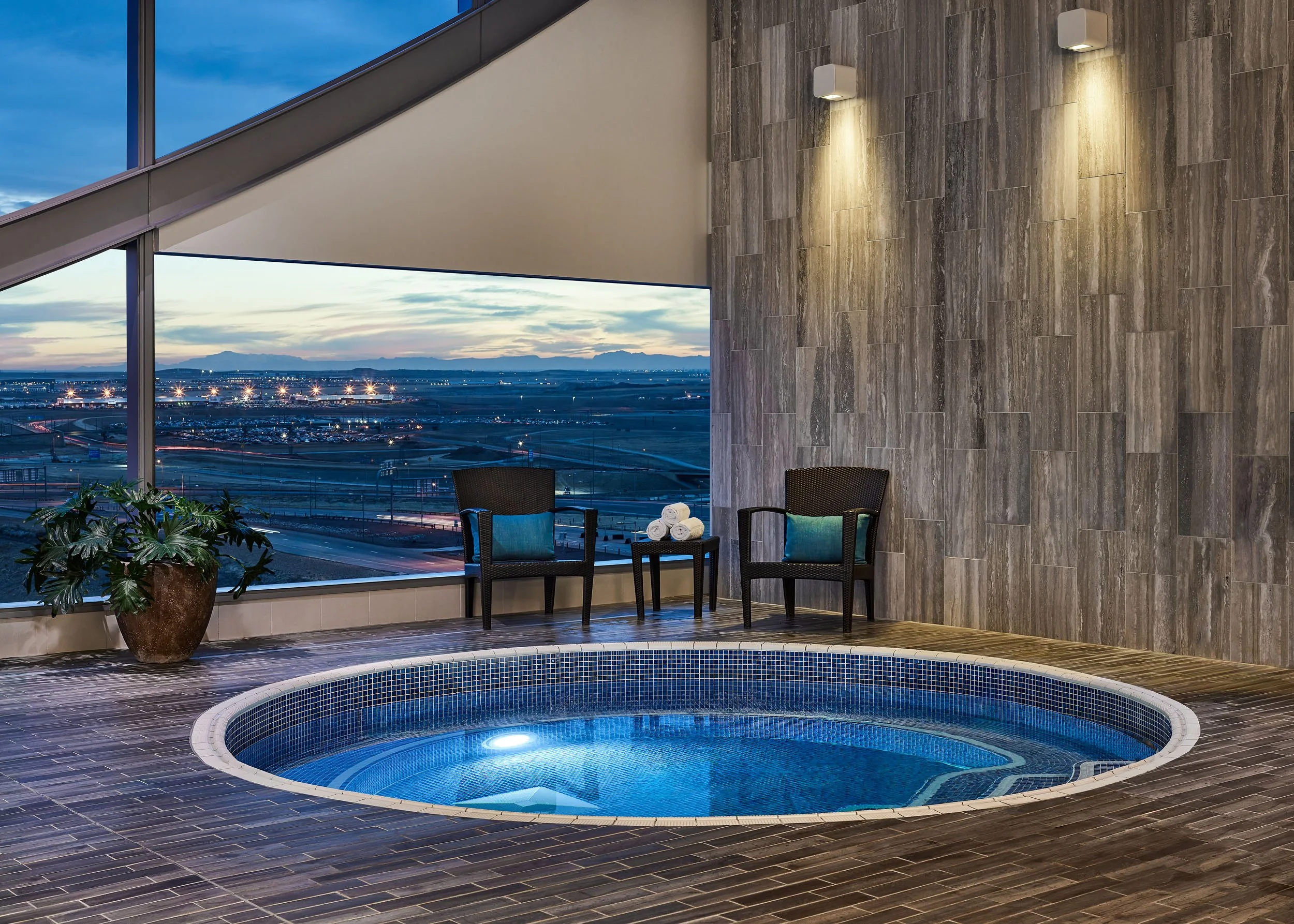 Indoor pool area with a hot tub, two chairs with cushions, a potted plant, large windows showing an evening cityscape with mountains in the distance, and modern wall-mounted lights.