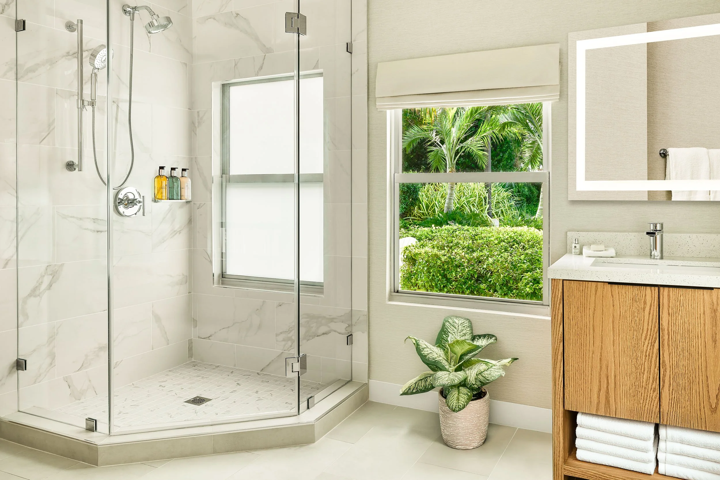 Modern bathroom with a glass-enclosed shower, white marble tiles, a window view of green tropical plants, a wooden vanity with folded white towels, and indoor plants.