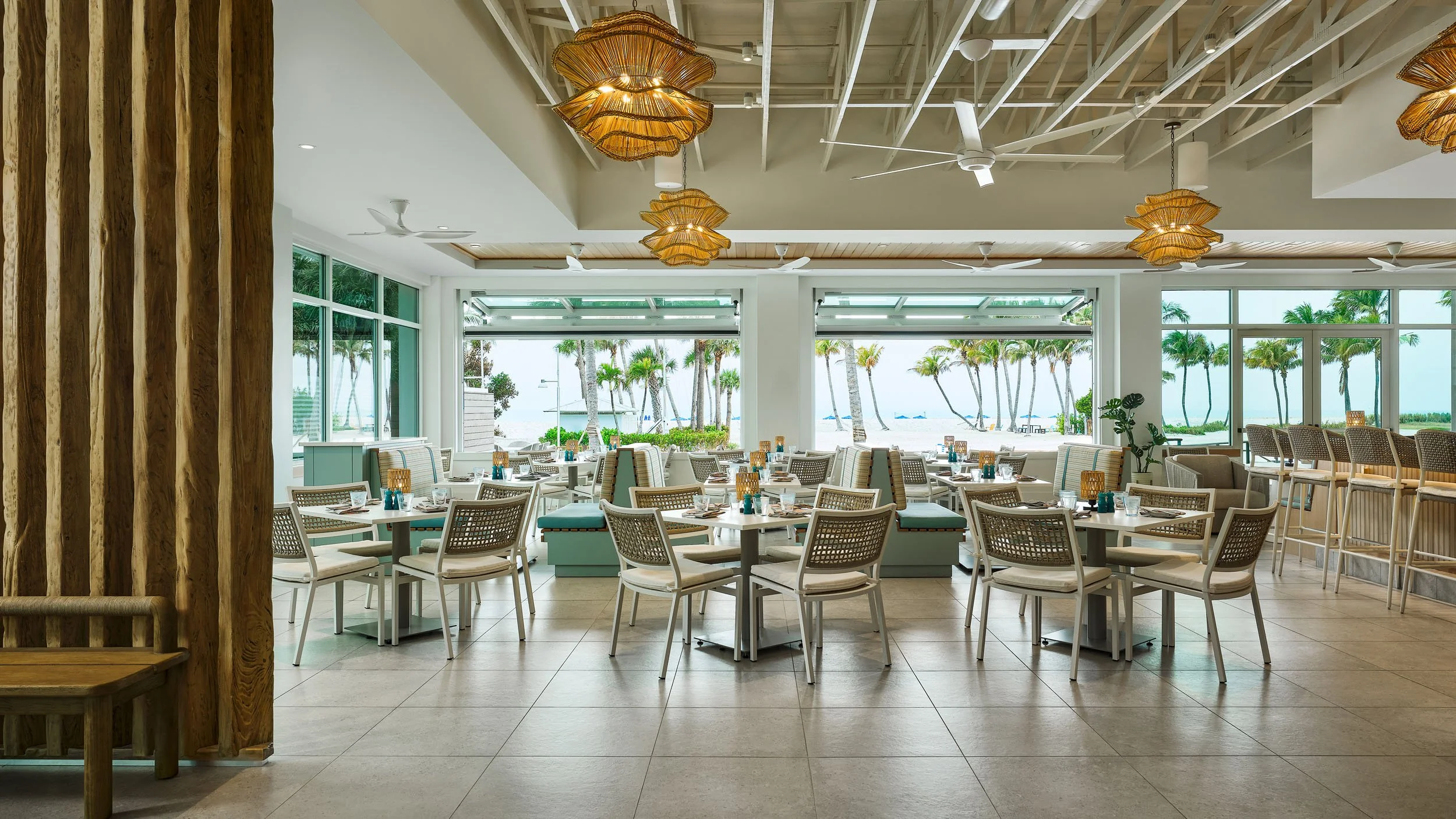 Bright beachside restaurant with white tables and chairs, large windows showing palm trees and the beach outside, and decorative woven pendant lights hanging from a white ceiling.