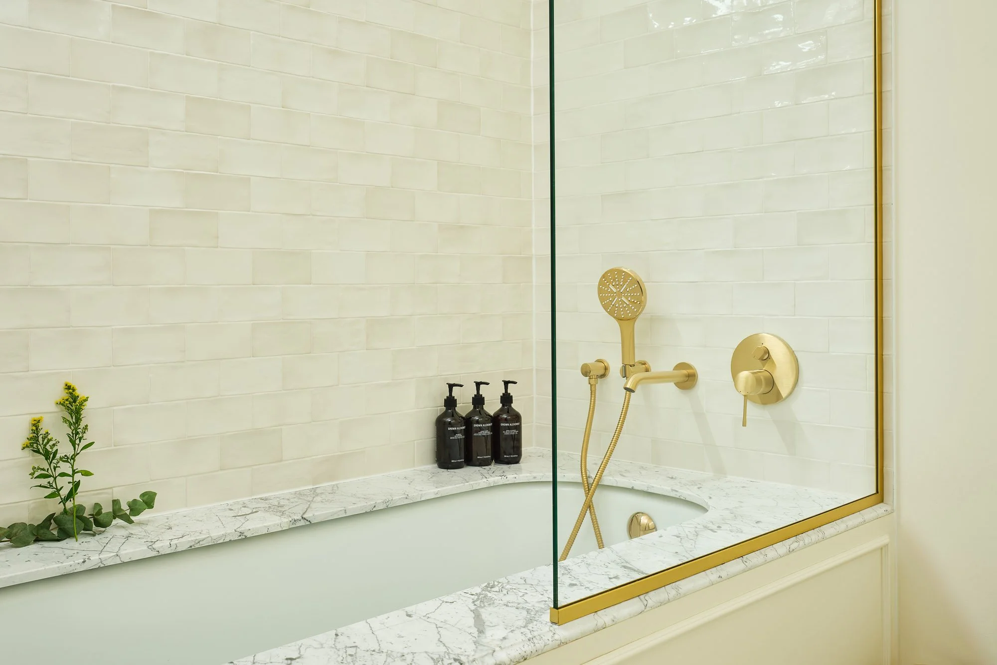 Modern shower with a beige tiled wall, a brass shower head, and matching fixtures. A marble shelf holds three black bottles, and a small plant is placed on the marble ledge below.
