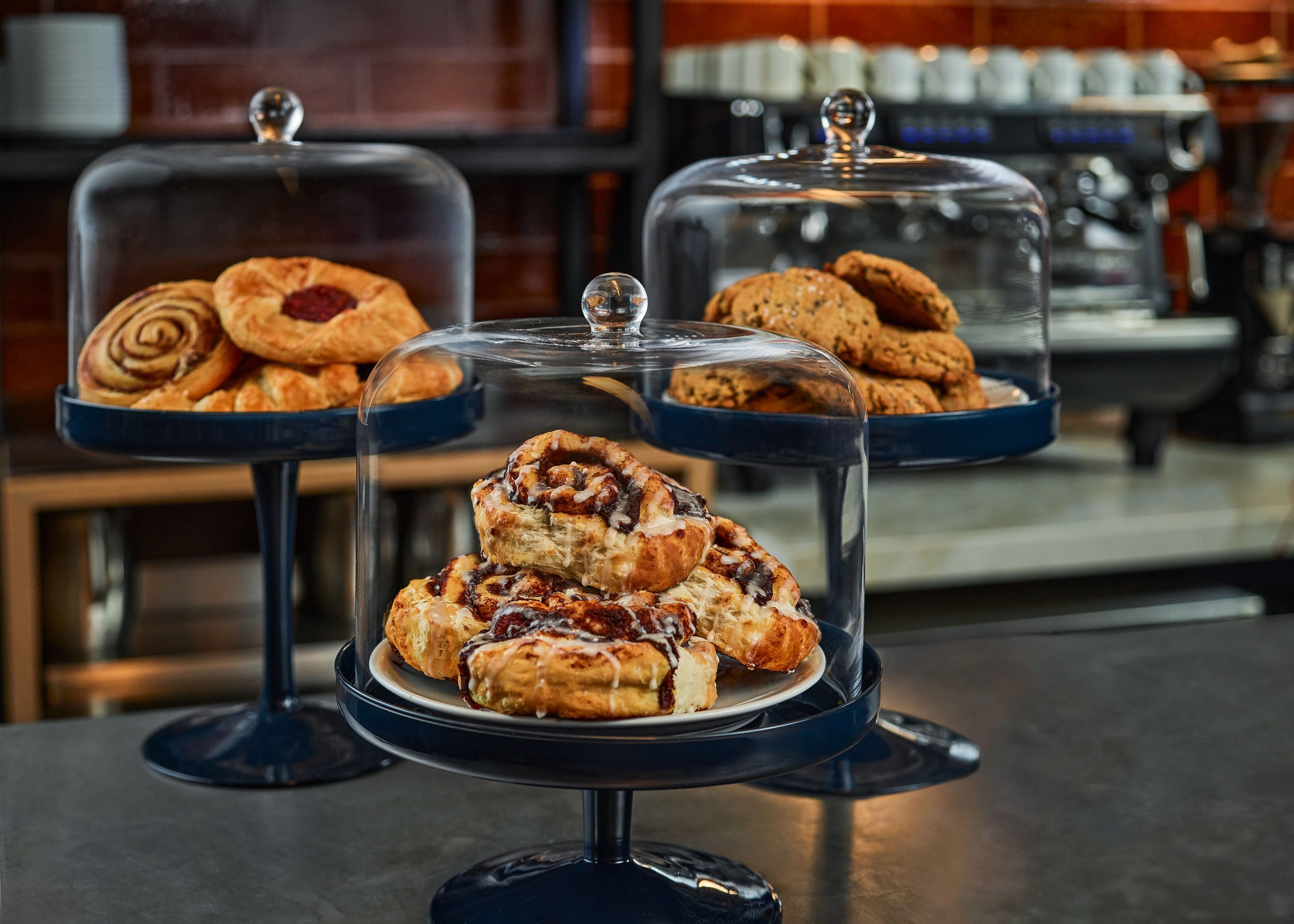 A three-tiered black dessert stand displaying a variety of cookies and pastries, with a glass cover on top of each tier. The background shows a brick wall and a coffee machine in what appears to be a cafe setting.
