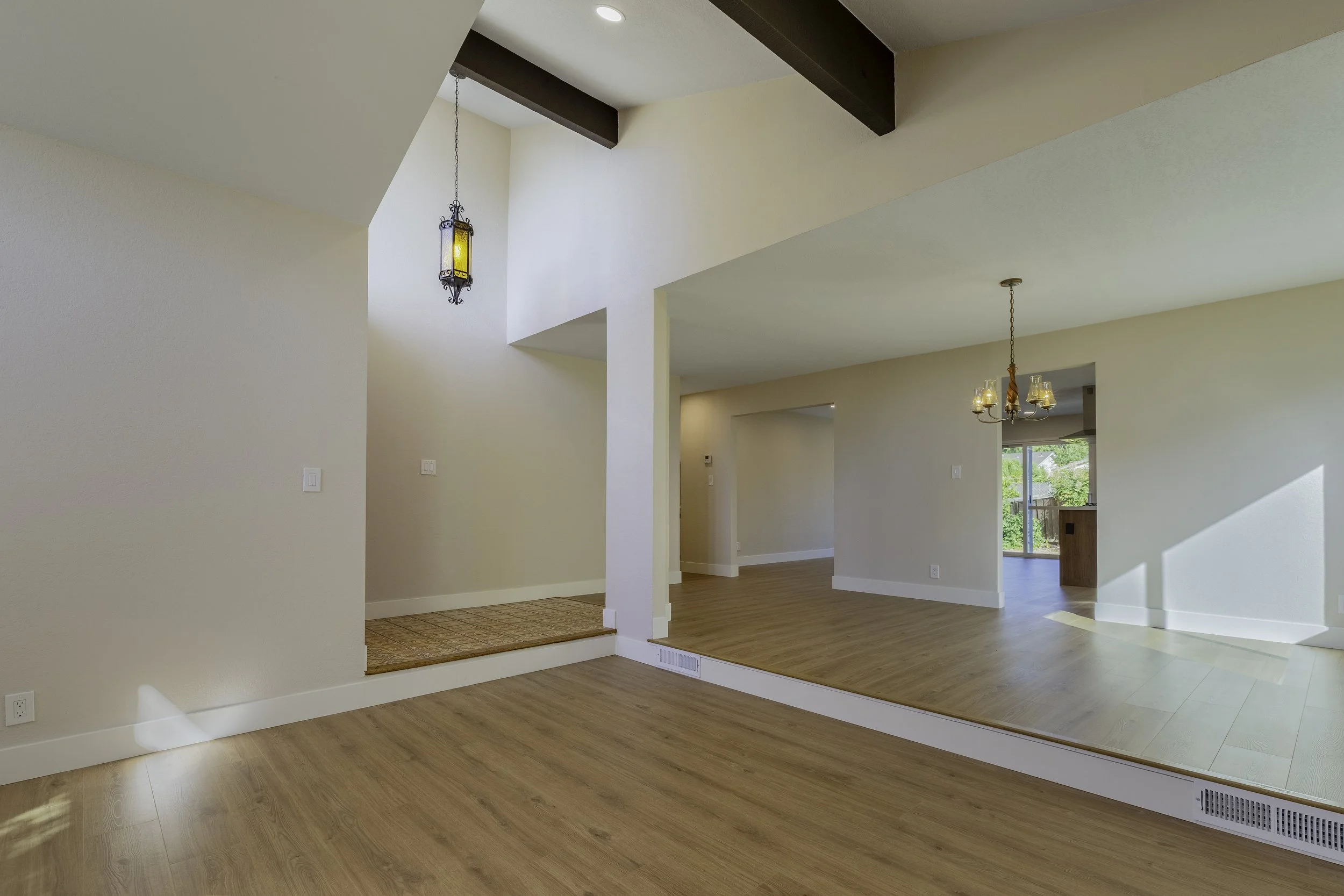 Open-concept living room with high ceiling, hardwood floors, and white walls, featuring a hanging chandelier and a decorative lantern-style ceiling light, with a view into the kitchen and outdoor patio.