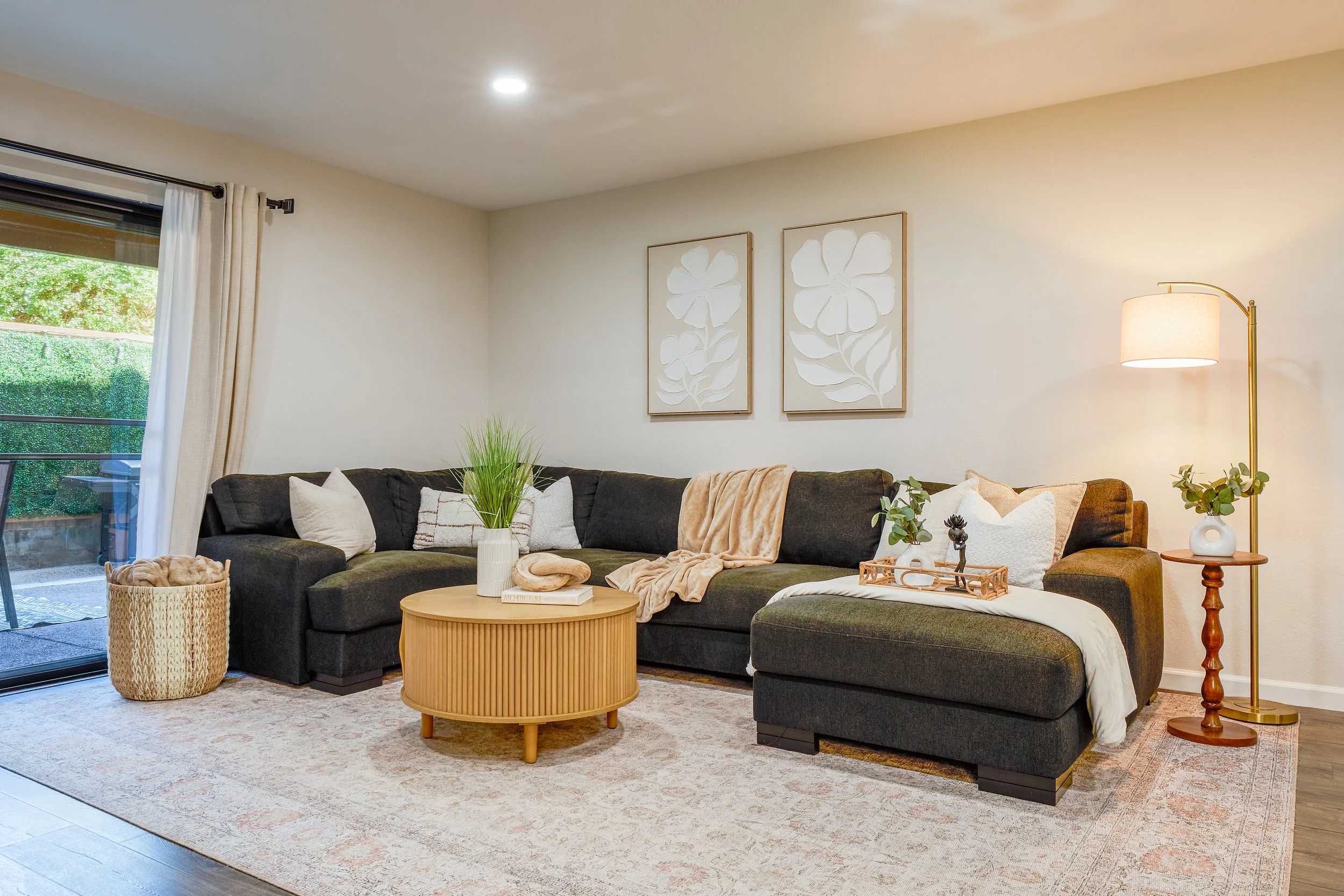 Living room with a dark gray sectional sofa, throw pillows, a round wooden coffee table, potted plants, wall art featuring white floral cutouts, a floor lamp with a white shade, a wicker basket, and sliding glass doors with white curtains.