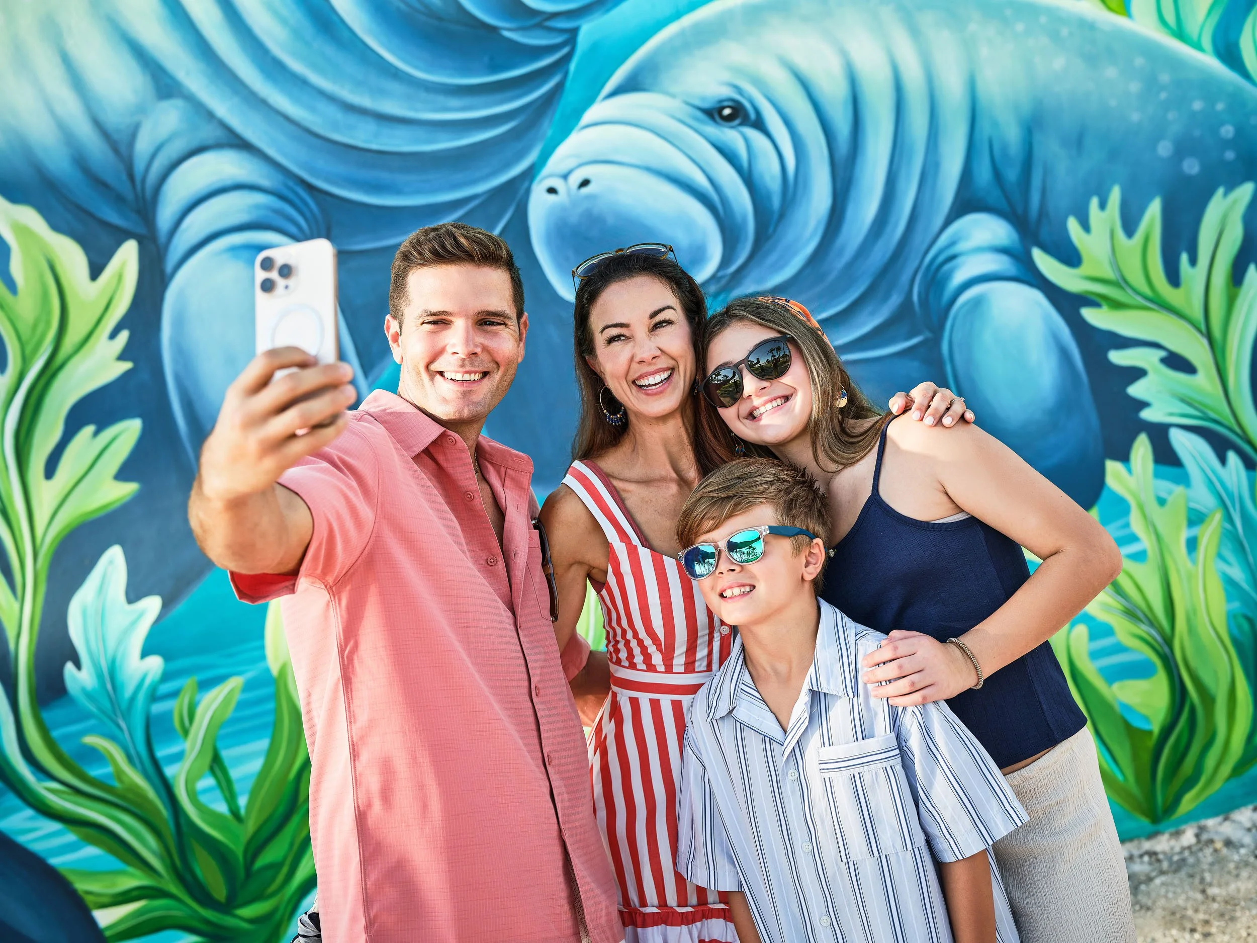 A smiling family of four taking a selfie in front of a colorful mural of a blue elephant with large green leaves on either side.