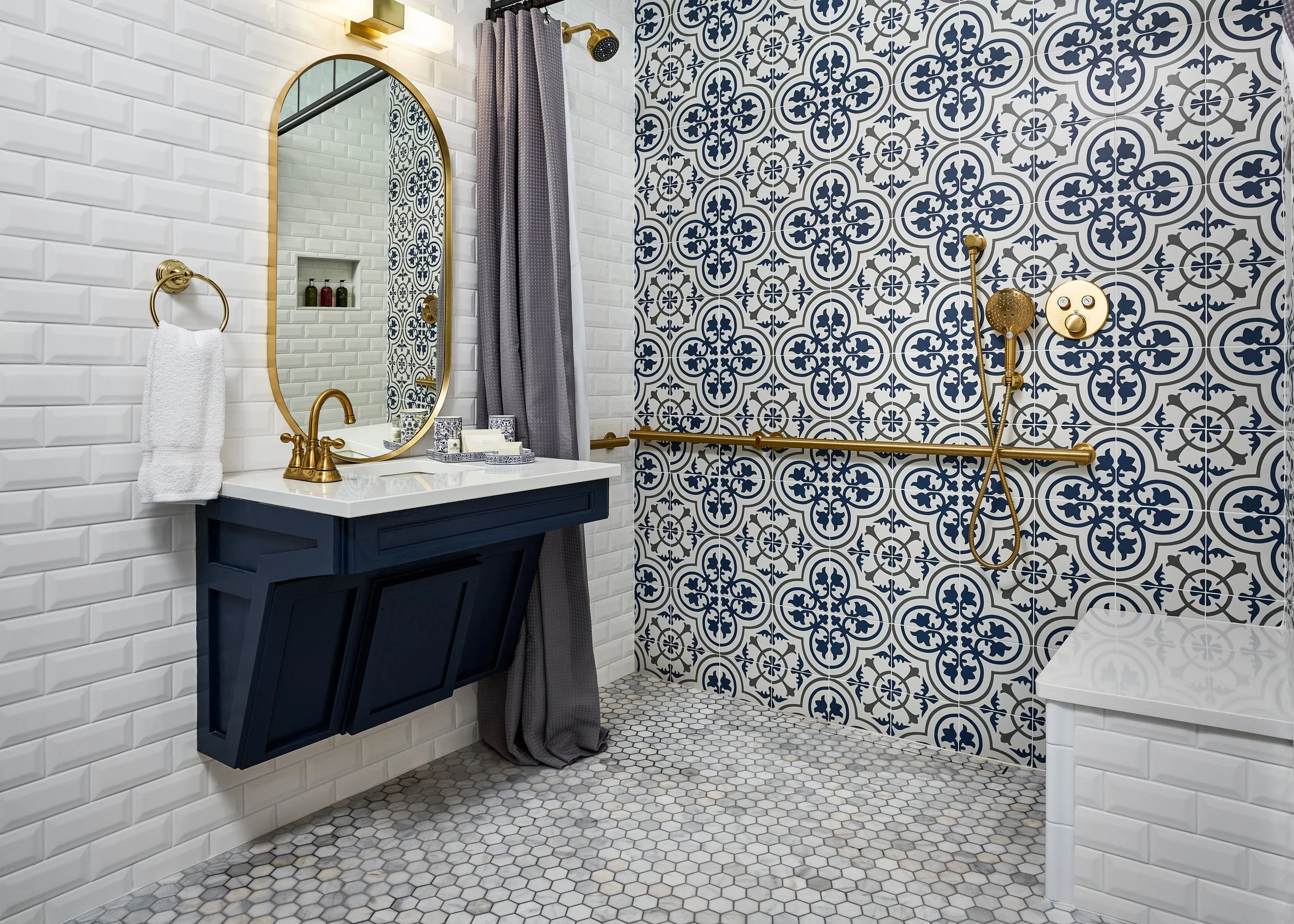 Bathroom with white subway tile on the left wall, decorative blue and white patterned tile on the shower wall, gold fixtures, a dark blue vanity with a white countertop, a round mirror, a white towel on a gold ring, and a gray shower curtain. The sho