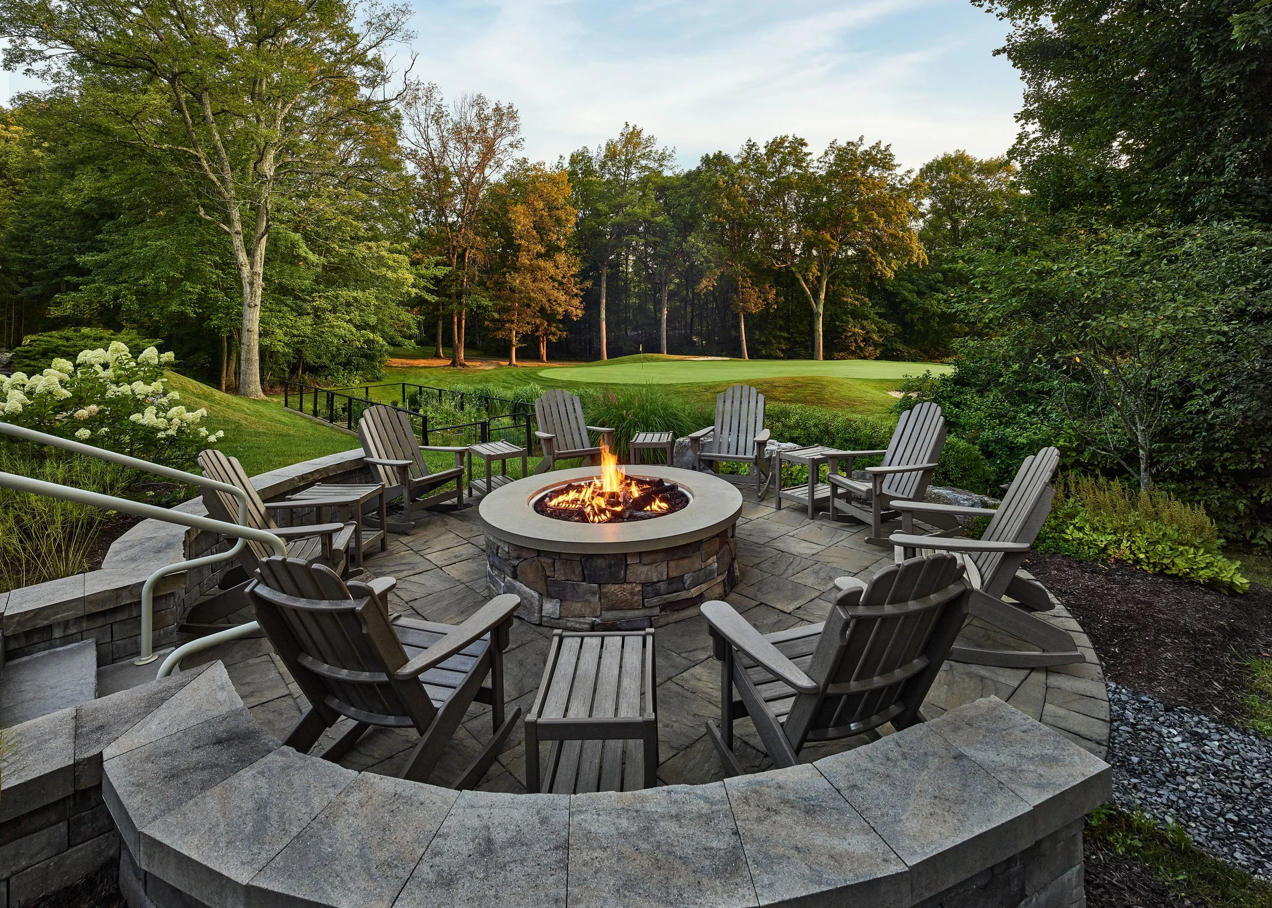 A backyard with a fire pit surrounded by Adirondack chairs, overlooking a golf course with green grass and tall trees.