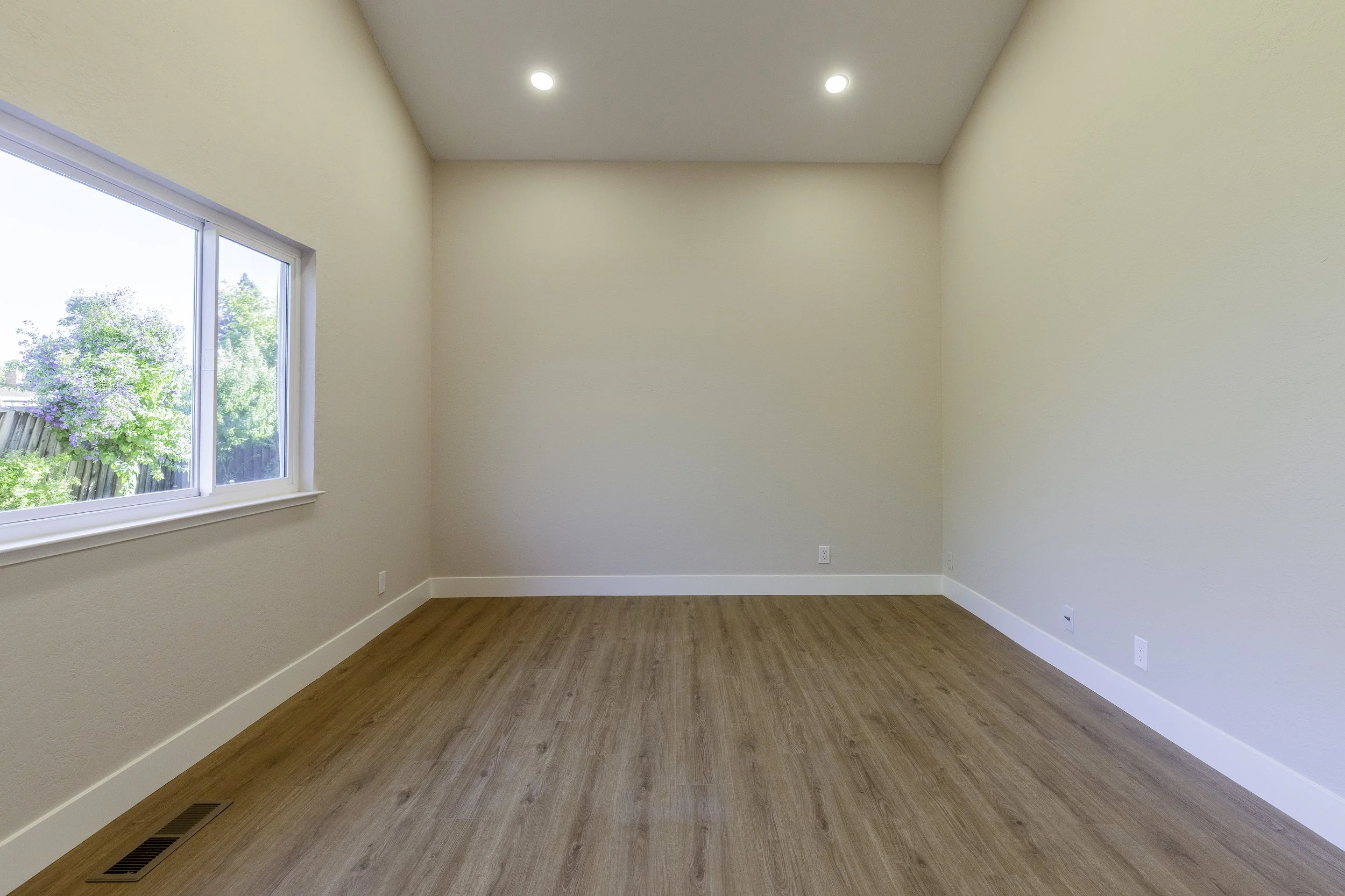 Empty room with a window showing trees outside, light-colored walls, wooden flooring, and recessed ceiling lights.