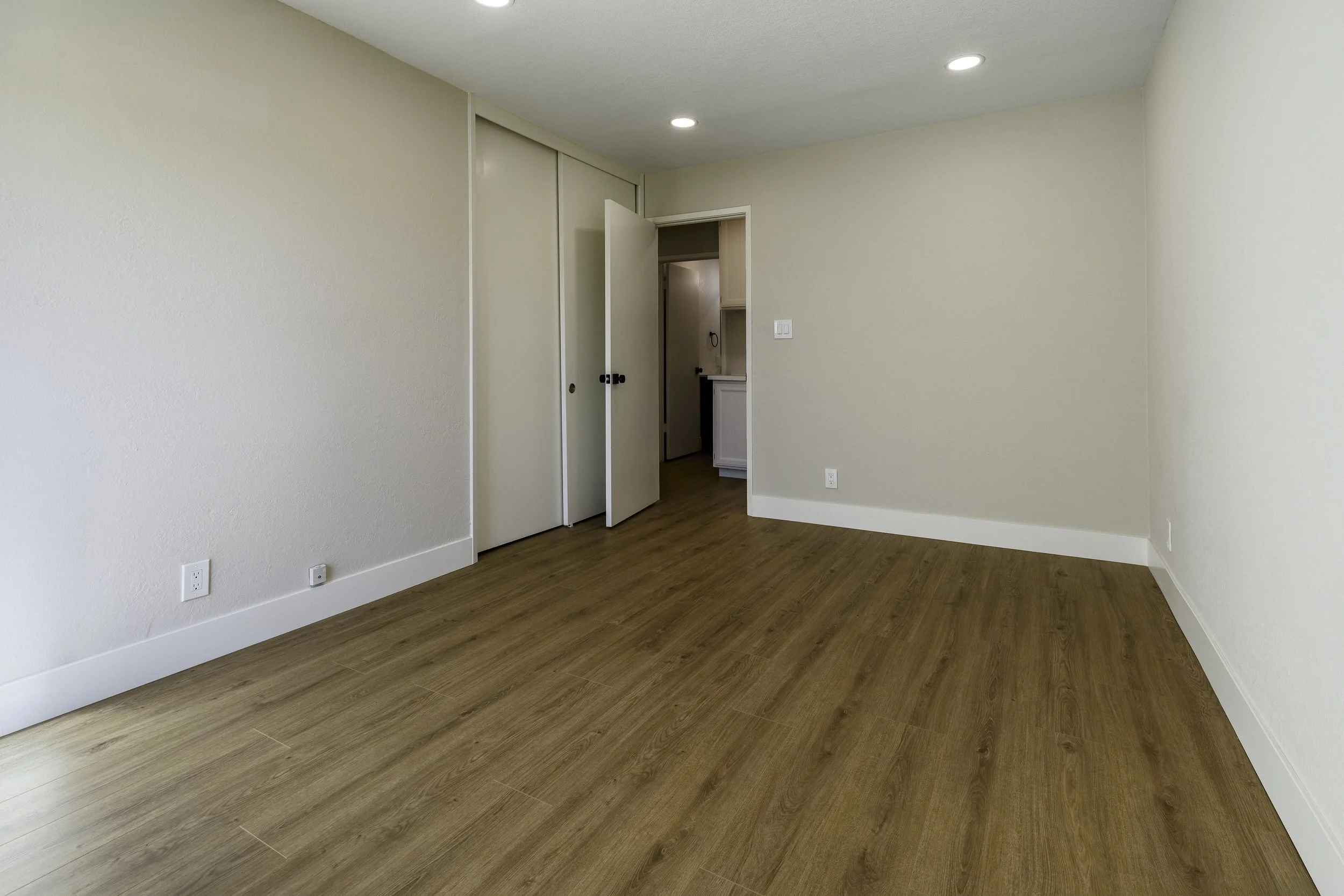 Empty room with white walls, wooden flooring, a closet with sliding doors, and a doorway leading to another room.