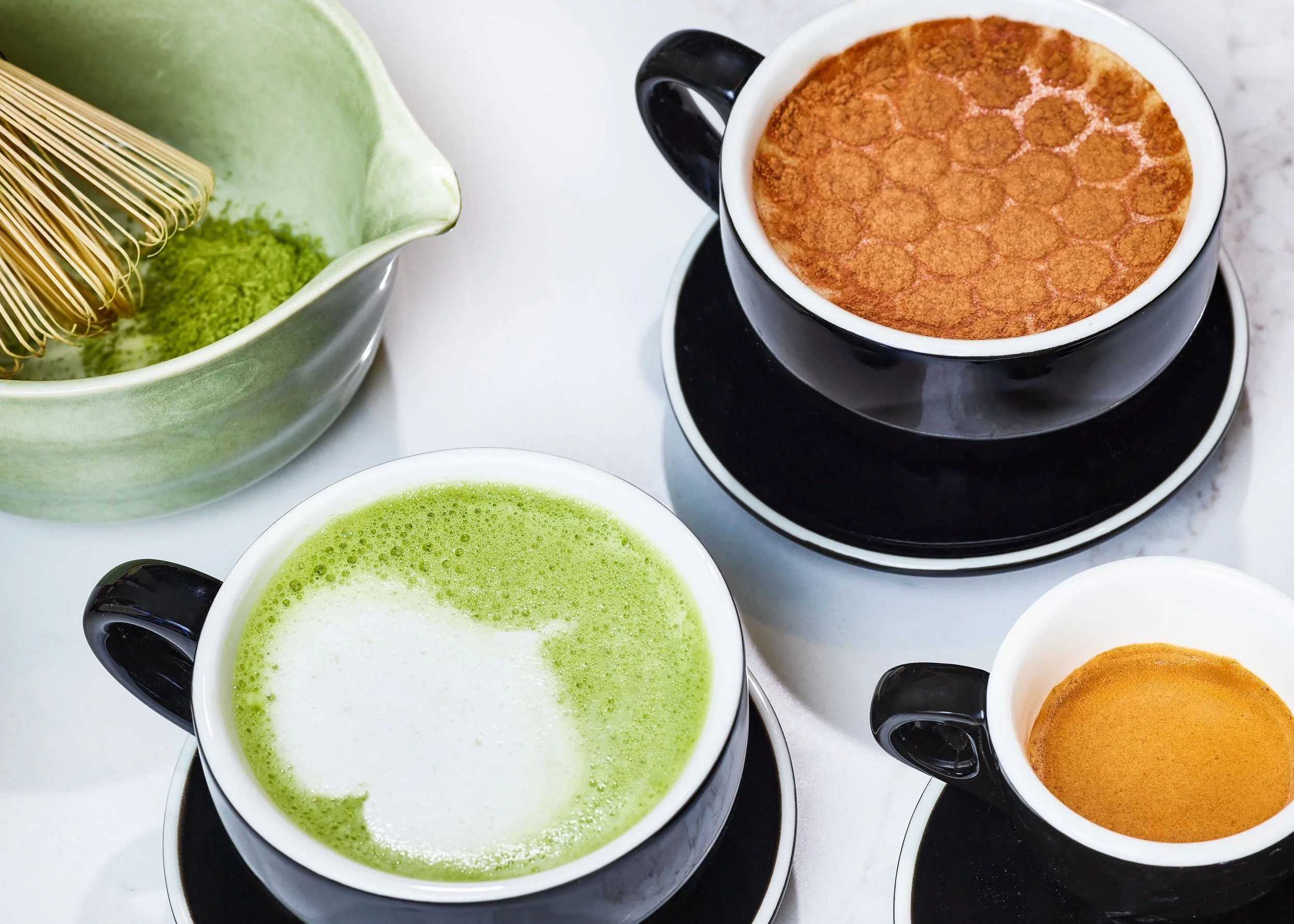 Close-up view of a white cup filled with a frothy green matcha latte, surrounded by other cups with various beverages, including a dark coffee drink and a frothy orange-colored drink, on a white surface.