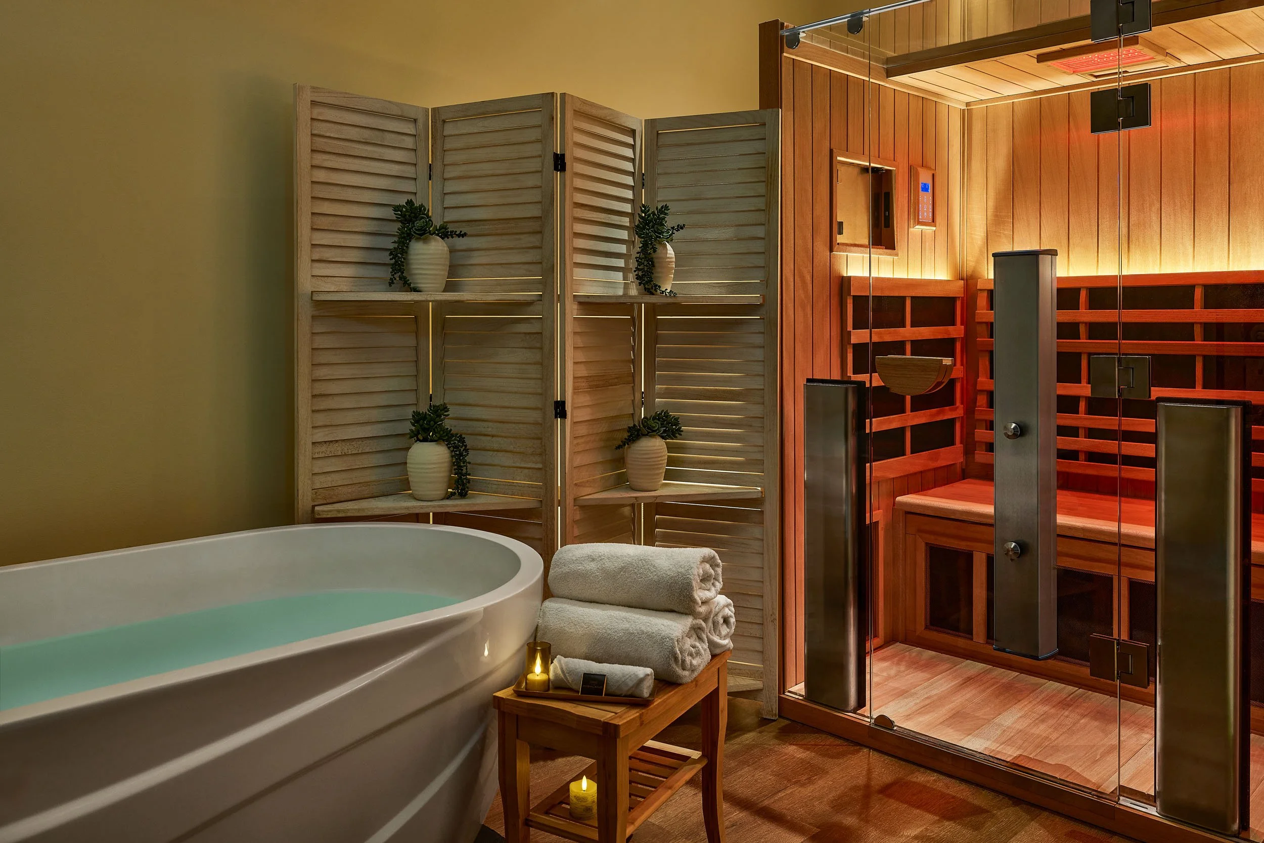 A spa room with a white bathtub filled with water, a wooden table with rolled white towels and candles, a wooden folding screen with vases and plants, and a sauna with wooden paneling and glass door.