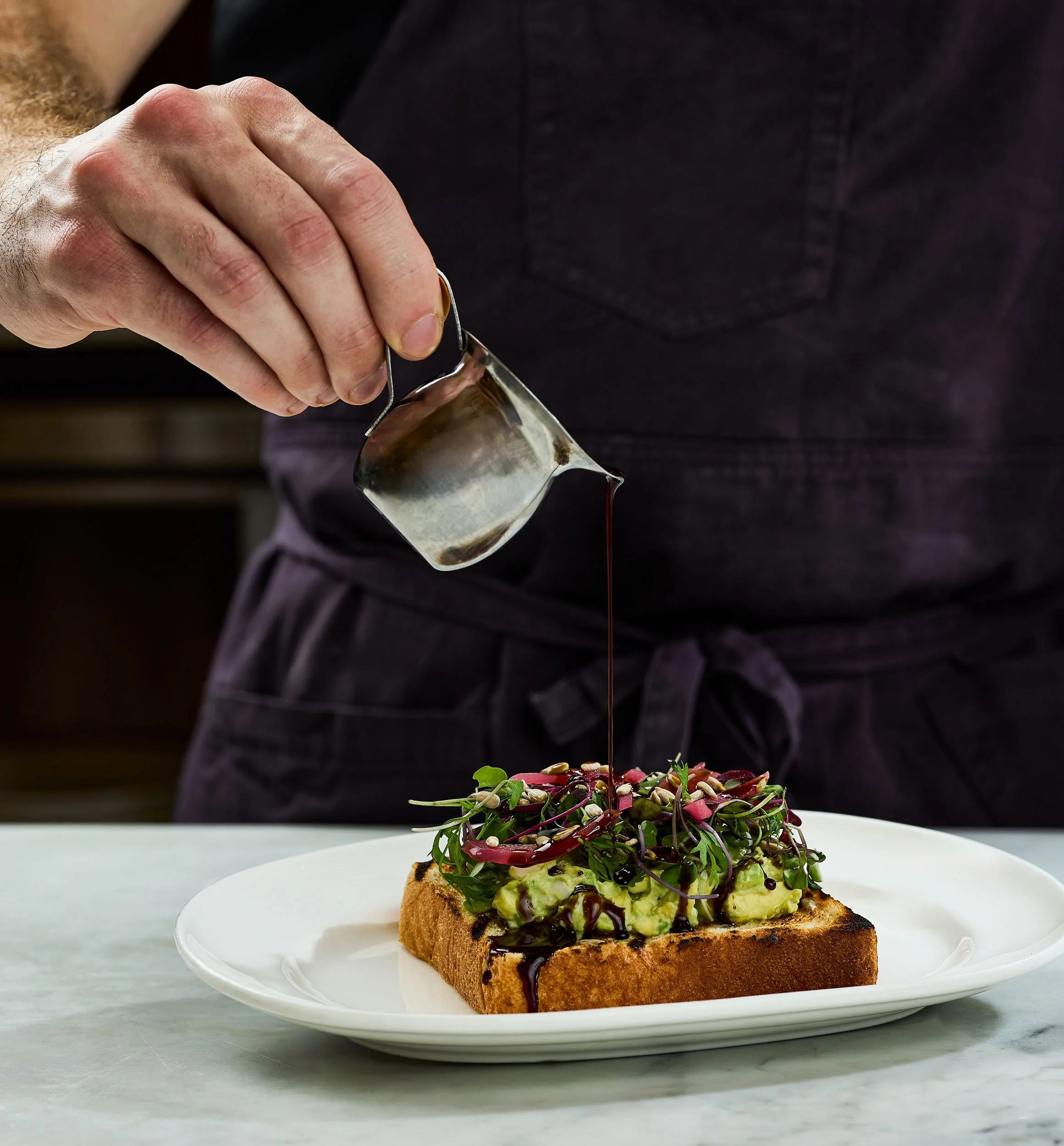 A person pouring balsamic glaze over an avocado toast topped with microgreens, seeds, and sliced radishes on a white plate.
