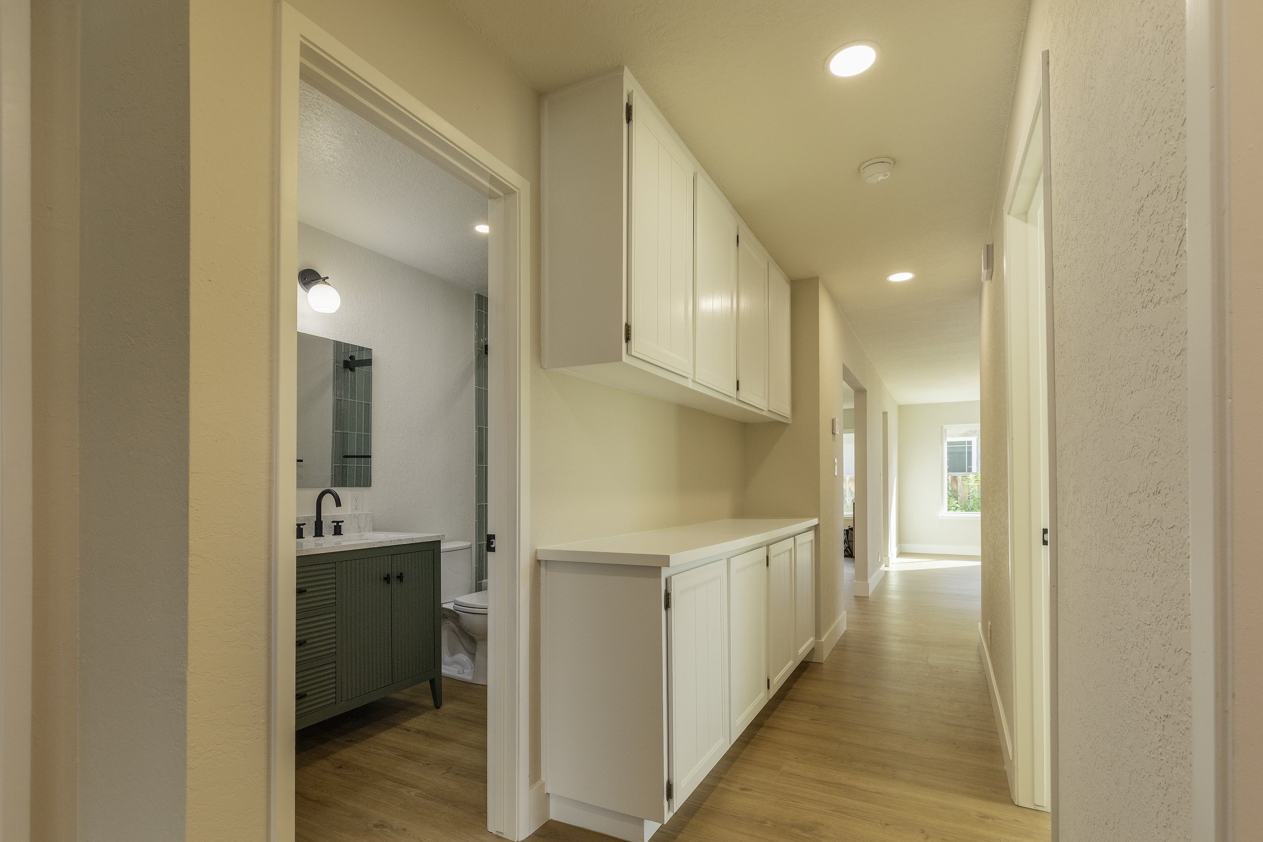 A hallway view of a modern home interior with beige walls and wooden flooring. To the left, a partial view of a bathroom with a green vanity and a mirror. On the right side, white cabinets are mounted on the wall, and in the distance, a window allows