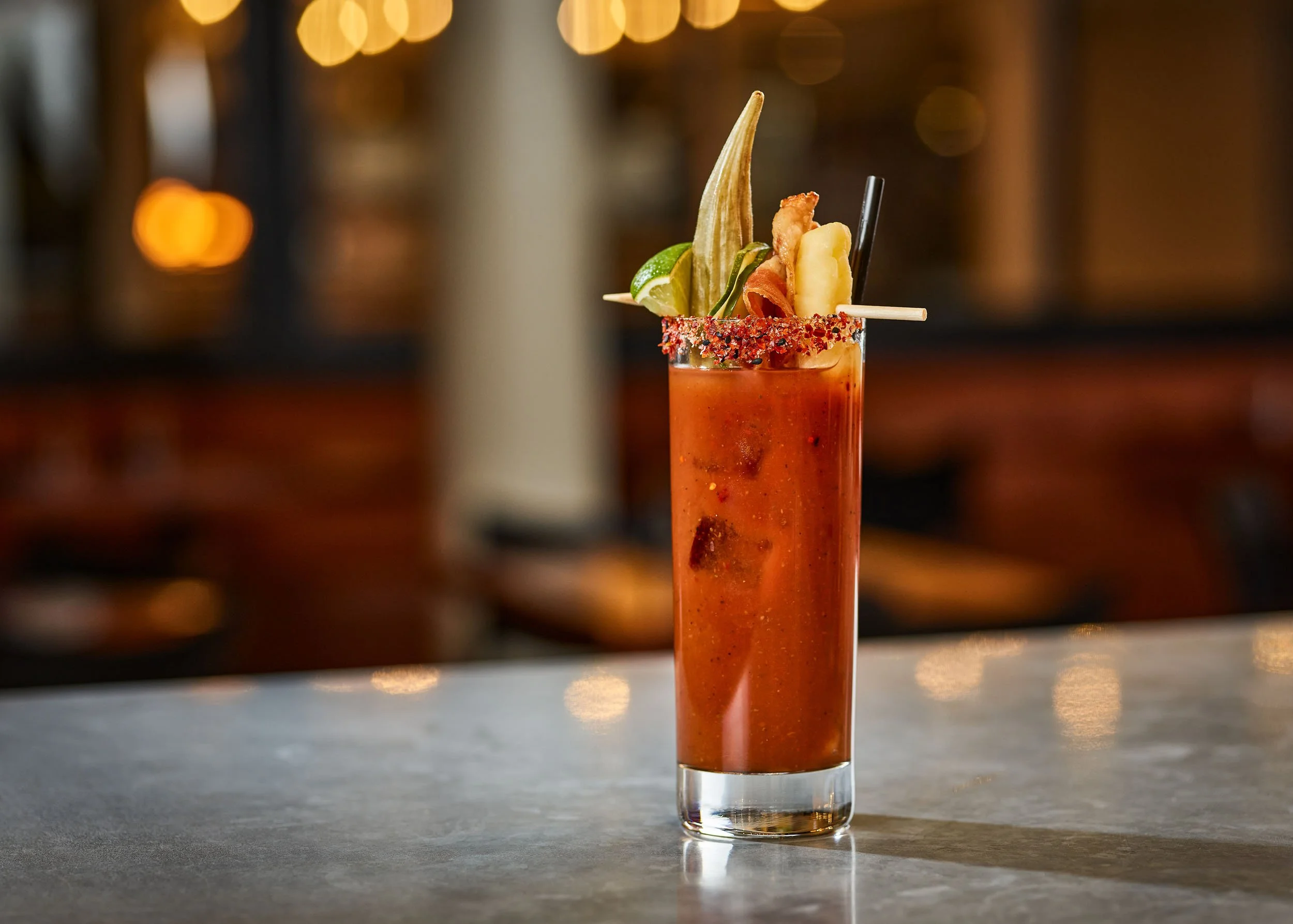 A tall glass filled with a spicy, red-colored mixed drink garnished with a lime wedge, a slice of banana, a piece of crispy bacon, a black straw, and a stick of dried banana chip, with a salted rim and blurred warm-toned bokeh lights in the backgroun