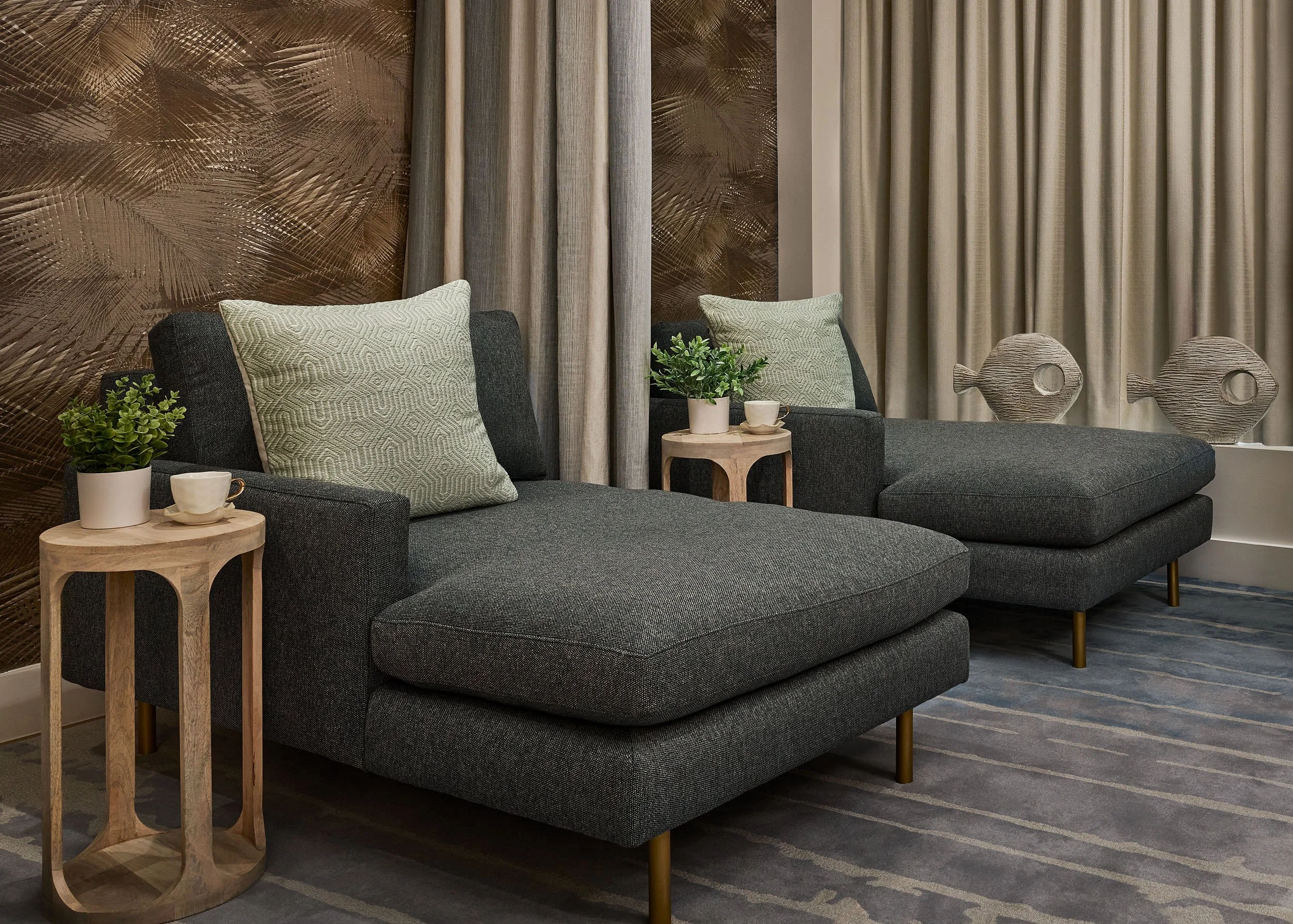 Living room with a dark gray sofa, light green pillows, small wooden side tables, potted plants, decorative fish sculptures, and curtains on the wall, with a textured brown wall behind.