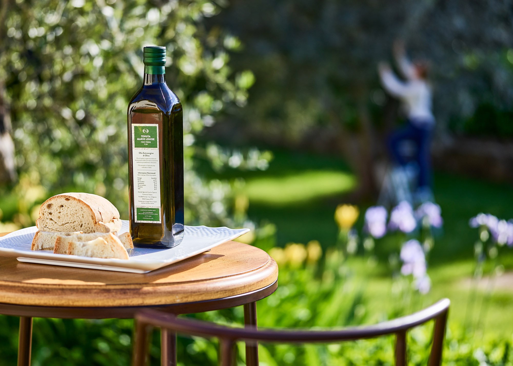 A white rectangular plate with slices of bread, a dark green bottle of olive oil with a label, on a round wooden table outdoors with green trees and a person in the background.