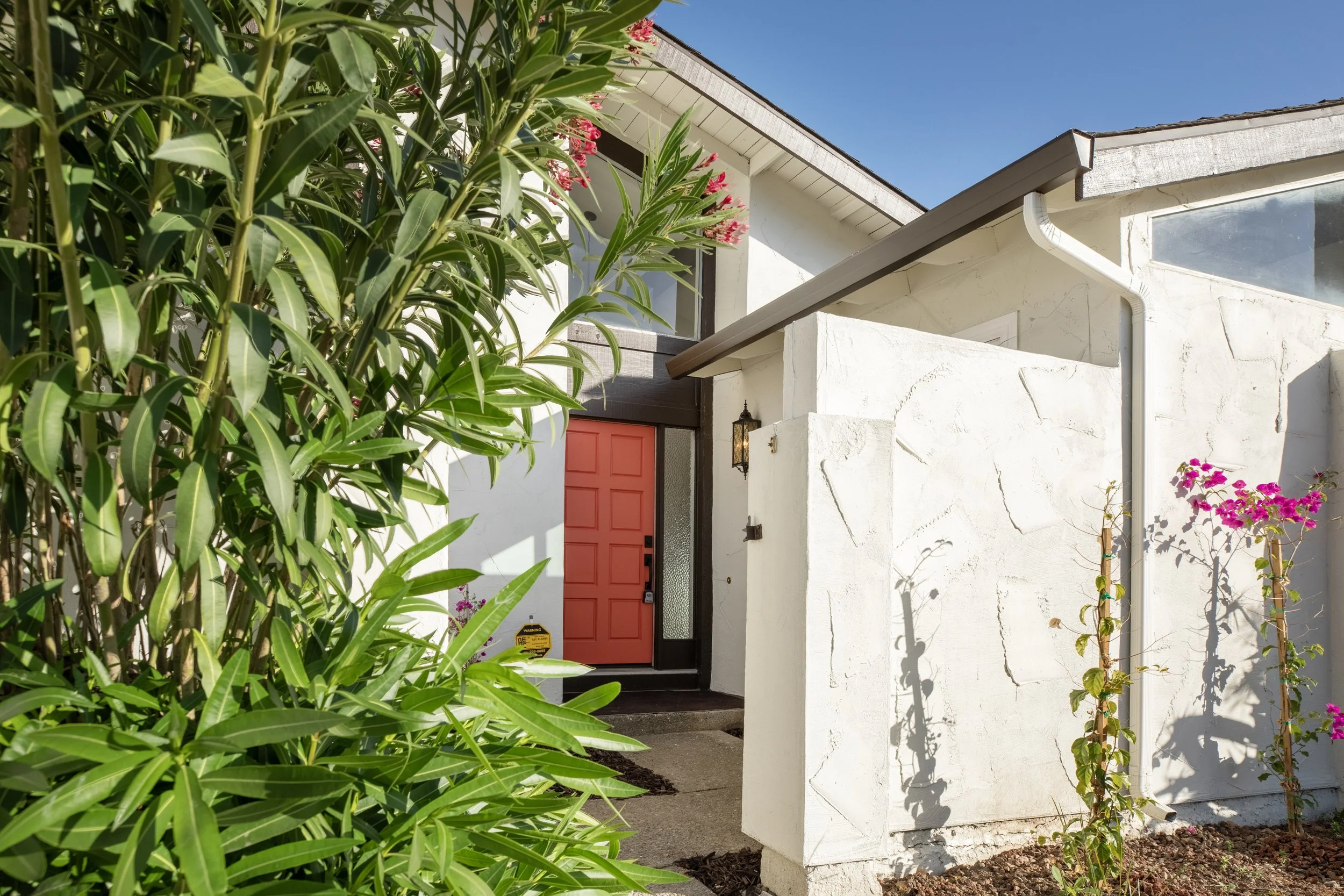 Front entrance of a house with a red door, surrounded by greenery and flowering plants, with a white exterior wall and a small stone step
