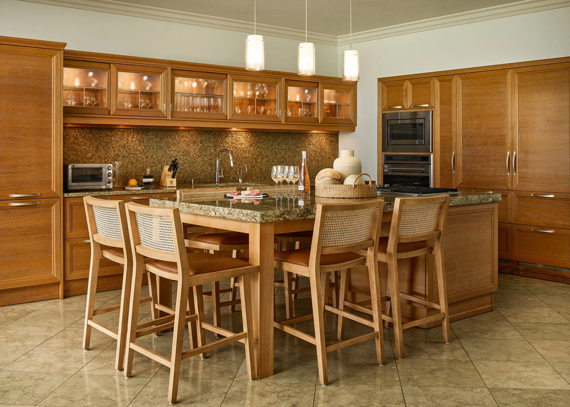 A kitchen with wooden cabinets, a granite countertop, and a kitchen island with chairs. There are glasses, a wine bottle, and decorative objects on the island. The lighting is warm with pendant lights hanging from the ceiling.