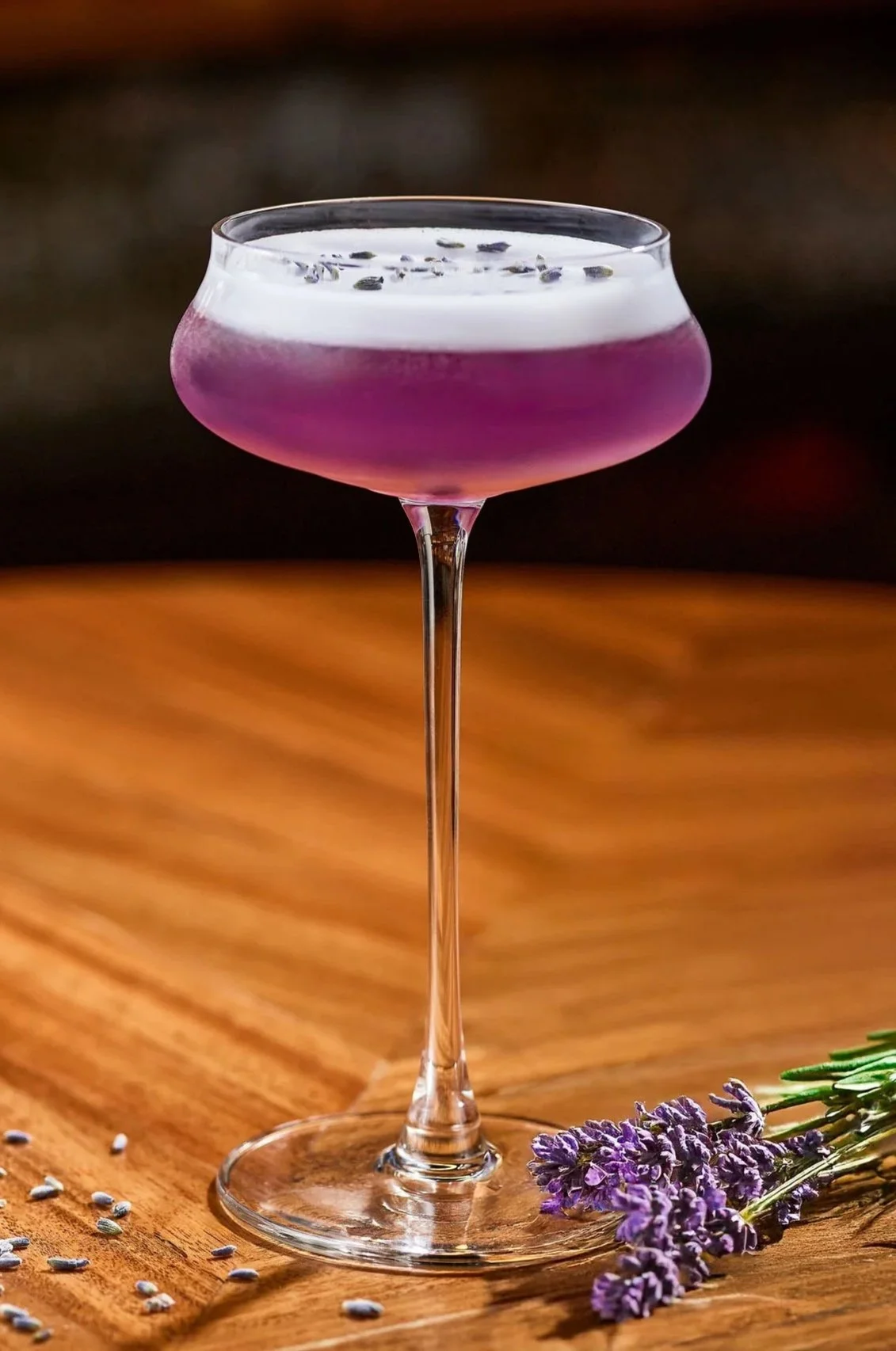 A cocktail in a stemmed glass with a purple hue, topped with foam and lavender buds, placed on a wooden surface with lavender sprigs and scattered lavender seeds.