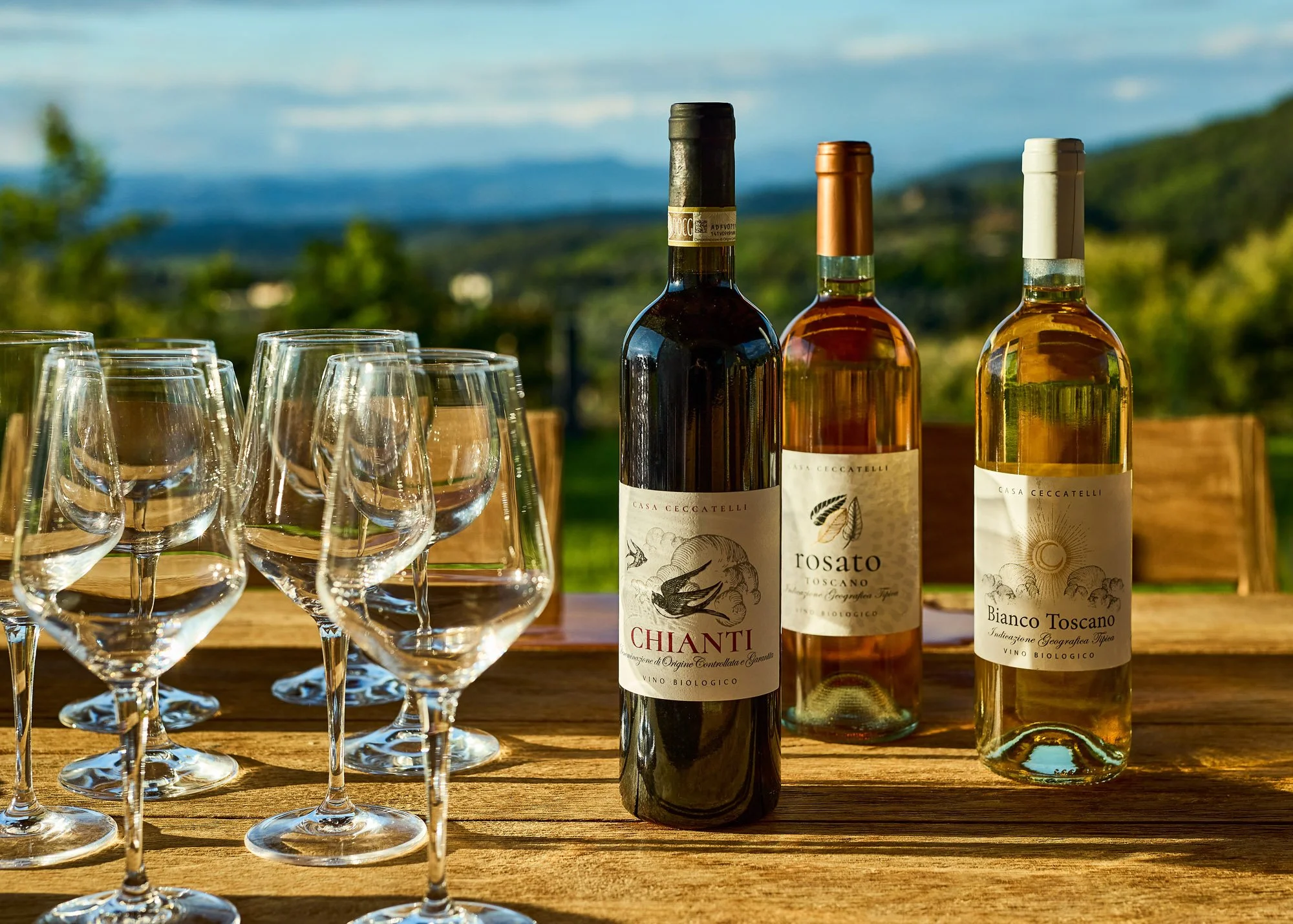 Three bottles of wine with labels, surrounded by multiple empty wine glasses on a wooden table outdoors with a scenic landscape of blue sky and green hills in the background.