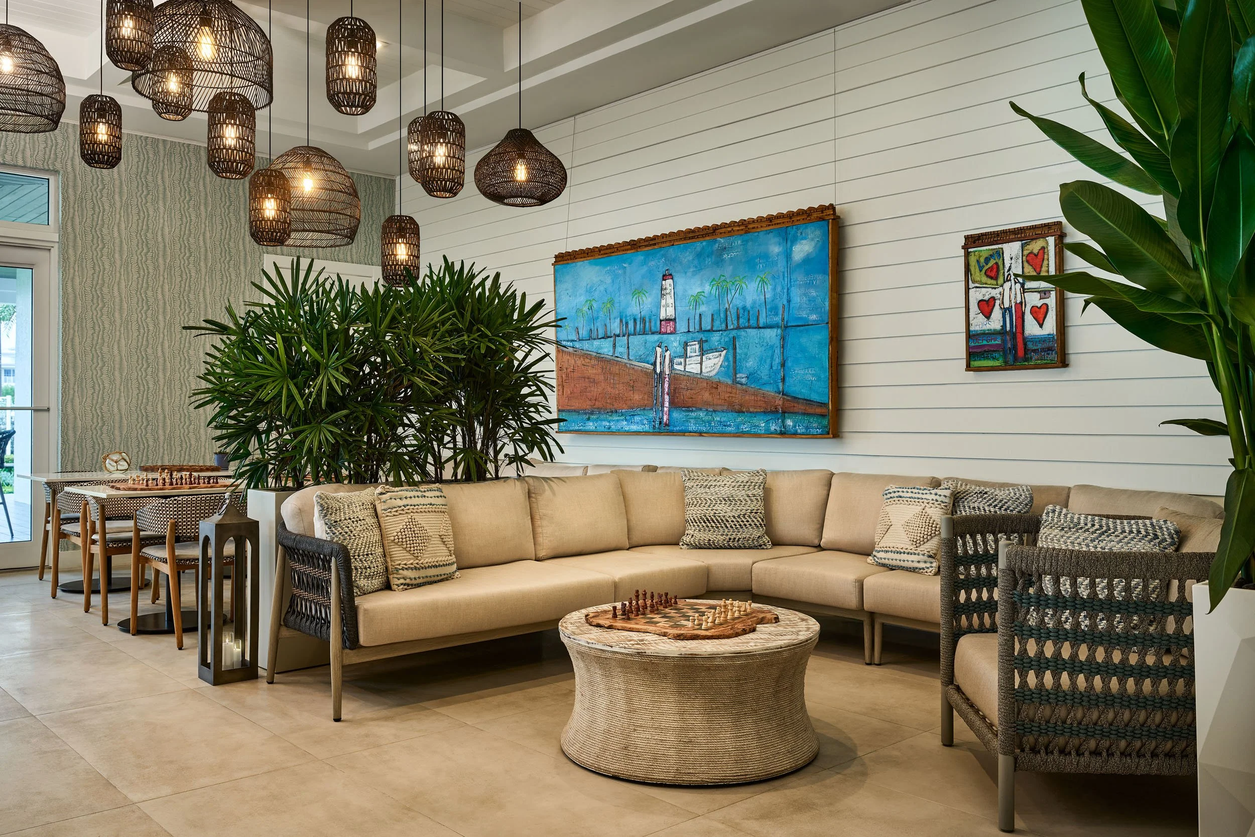 Interior of a living room with a large L-shaped beige sofa, decorative pillows, a woven coffee table, and green plants. On the wall, there are two colorful paintings, one depicting a lighthouse and boats, and the other with hearts and a person. Hangi