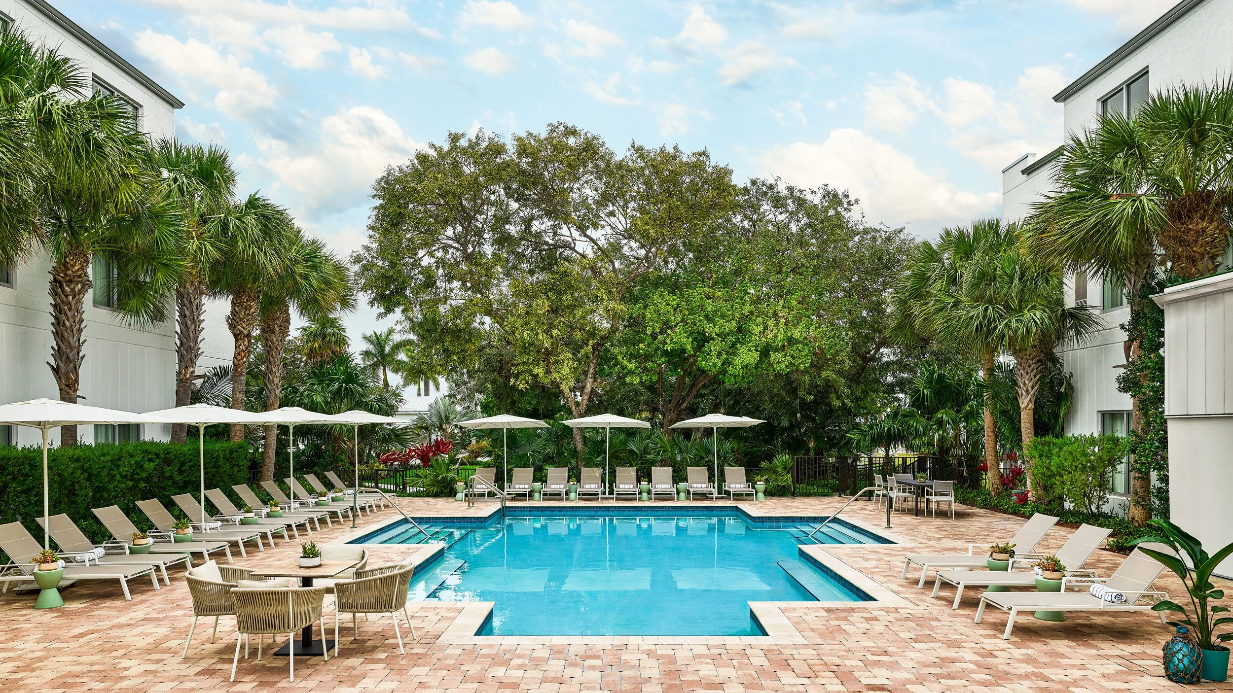 Swimming pool area with lounge chairs and umbrellas, surrounded by palm trees and greenery, in an apartment or hotel complex.