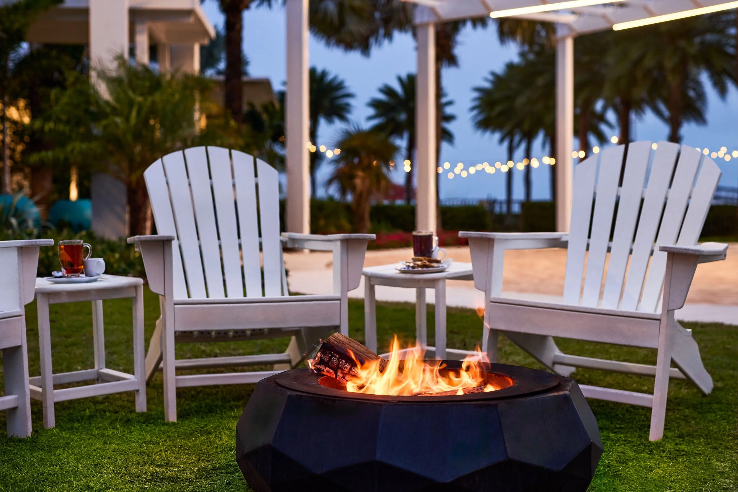 White Adirondack chairs arranged around a fire pit with a small fire burning, set outdoors with palm trees, string lights, and a building in the background at dusk.