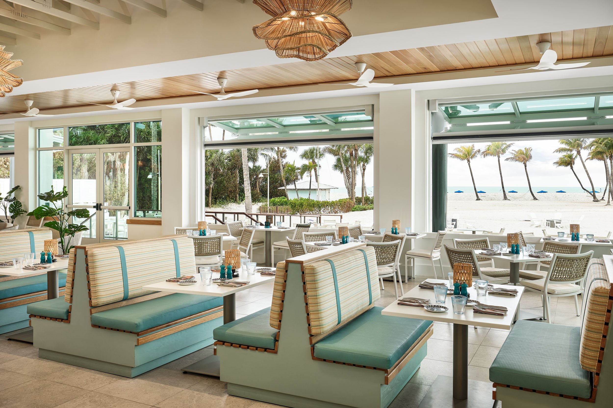 The image shows the interior of a beachside restaurant with white walls, a wooden ceiling, and large windows. The windows offer a view of the beach with sand, palm trees, and lounge chairs with umbrellas outside. Inside, there are tables set for dini