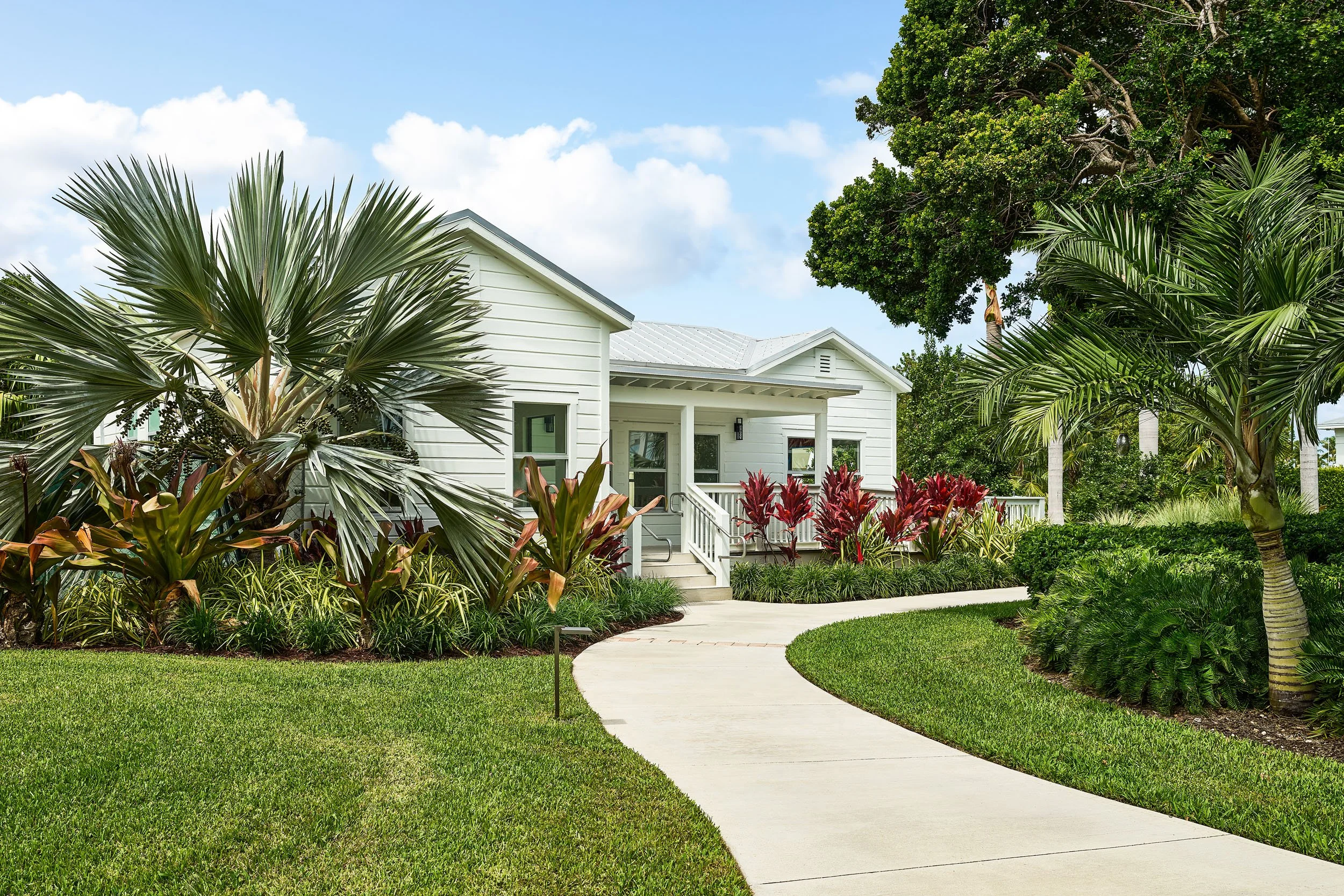 A white house with a porch, surrounded by lush green palm trees and tropical plants, with a curved concrete pathway leading to the entrance on a sunny day with blue sky and fluffy clouds.