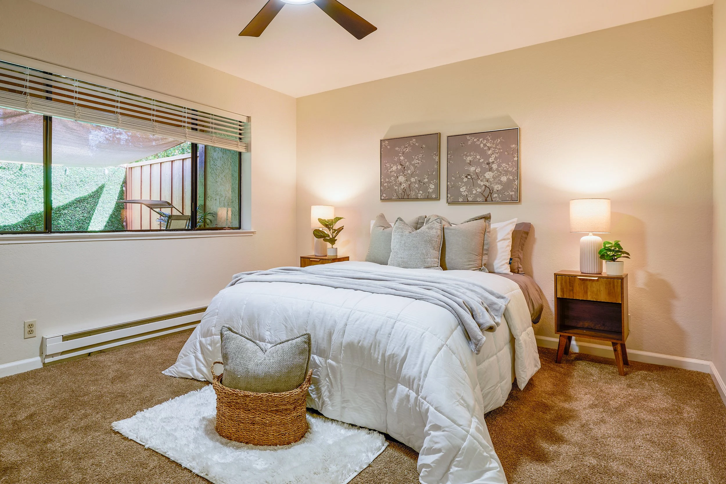 A cozy bedroom with a large bed, multiple pillows, bedside tables with lamps, and framed artwork on the wall. There is a window with blinds and a view of greenery outside. The room features beige walls, a ceiling fan, and a plush white rug under the bed.