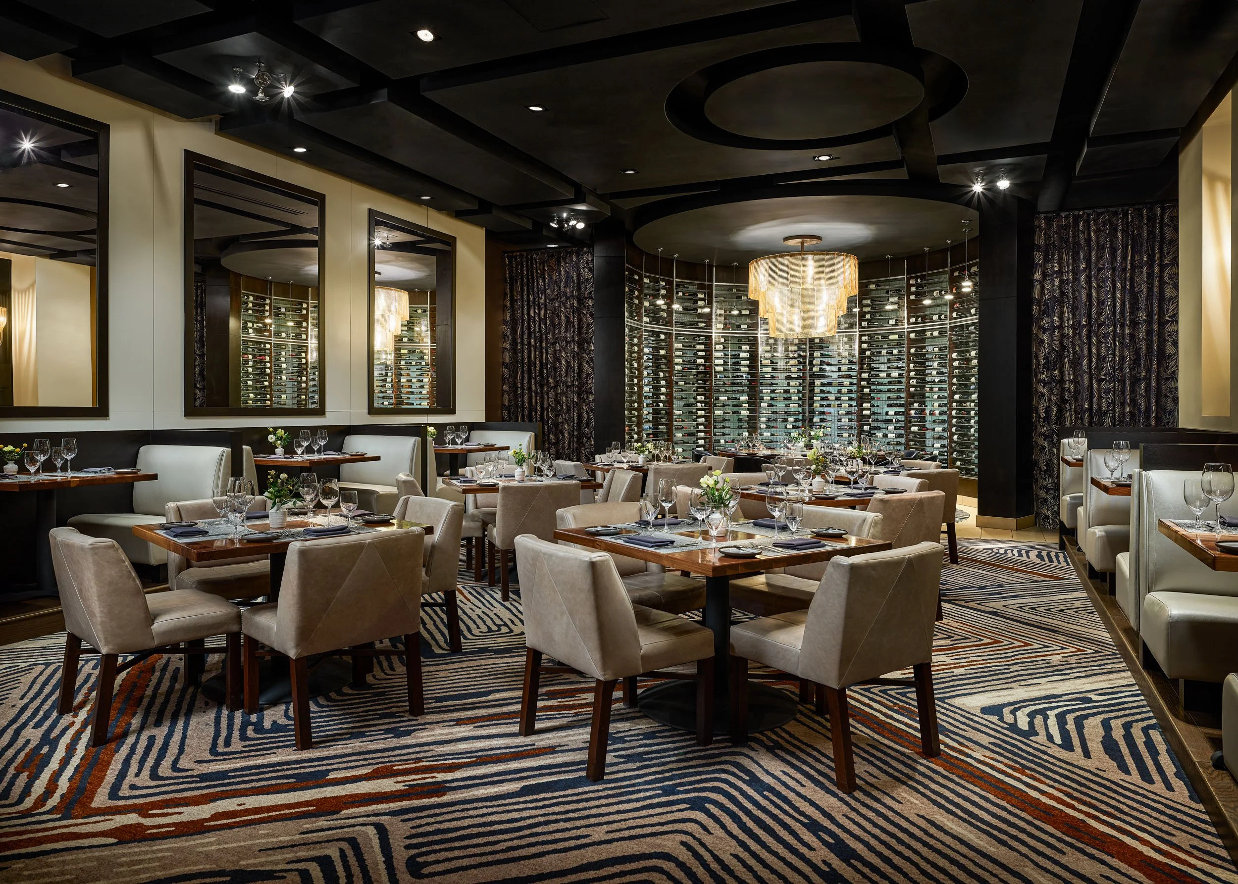 Elegant restaurant interior with beige upholstered chairs, tables set with wine glasses and cutlery, and a decorative wine wall with a chandelier overhead.
