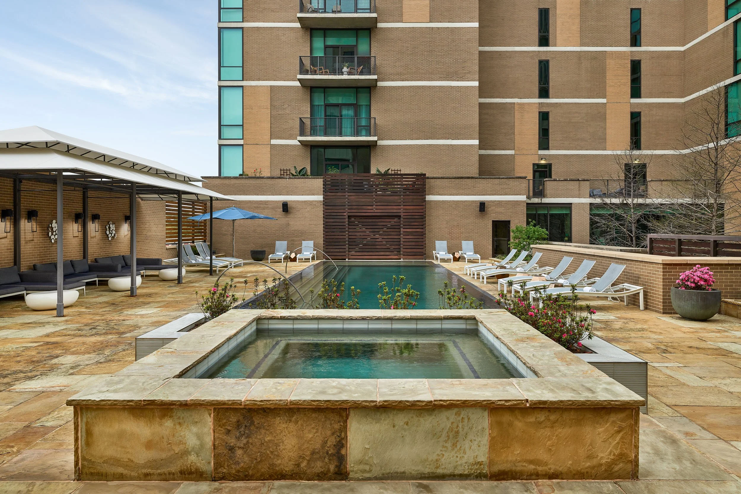 A modern apartment building's outdoor pool area with lounge chairs, umbrellas, and greenery.