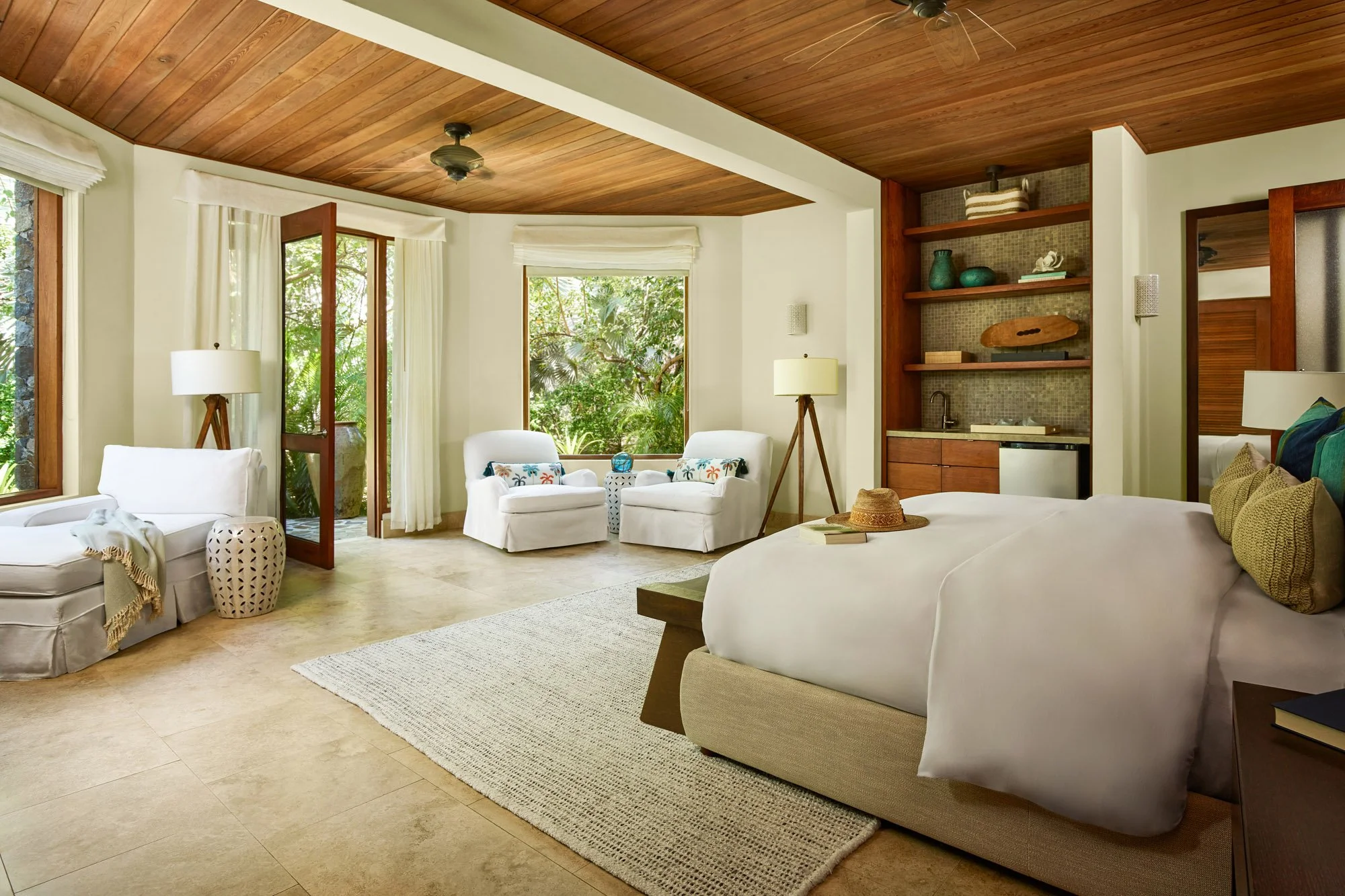 A spacious, bright living room with a wooden ceiling, large windows with white curtains, and a view of outdoor greenery. Furniture includes a white sofa, white armchairs with decorative pillows, a standing lamp, and a small side table. A built-in she
