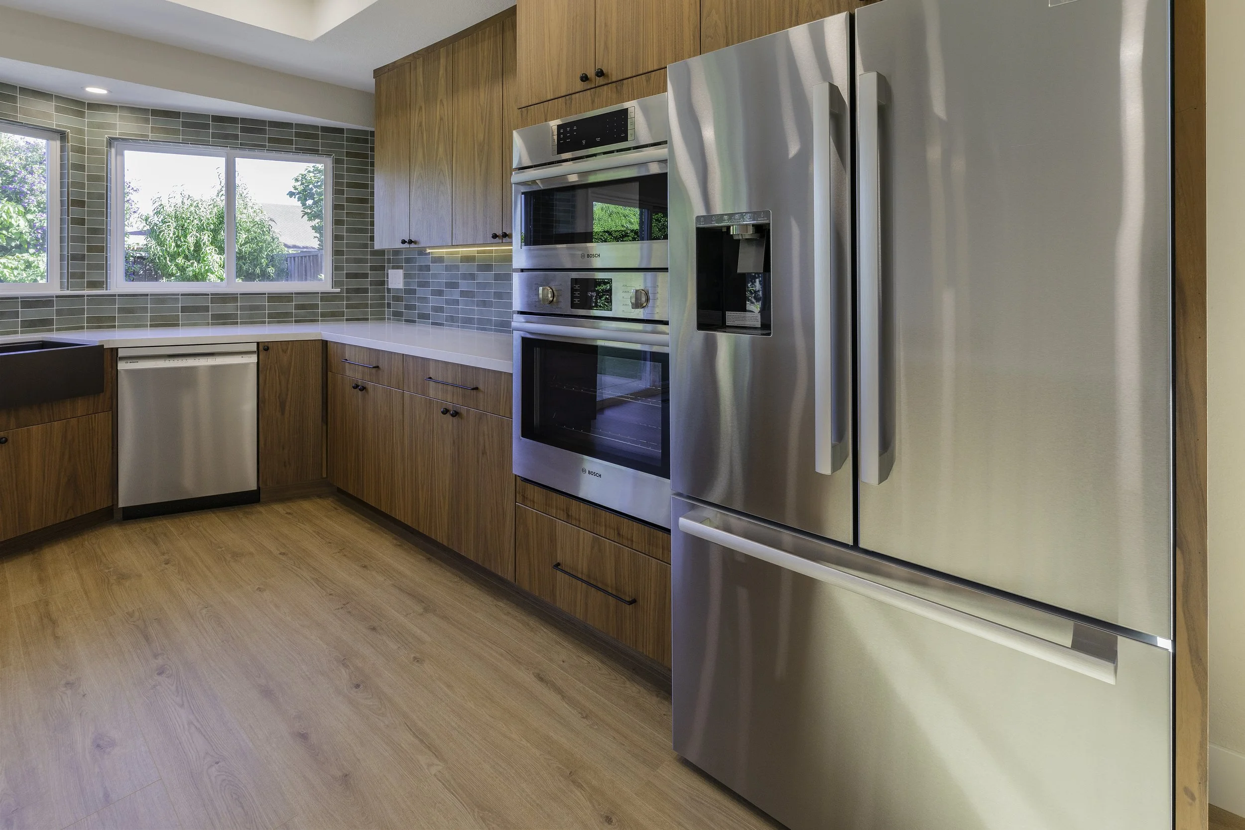 Modern kitchen with wooden cabinets, stainless steel appliances, large window showing green outdoor trees, and light-colored wood flooring.