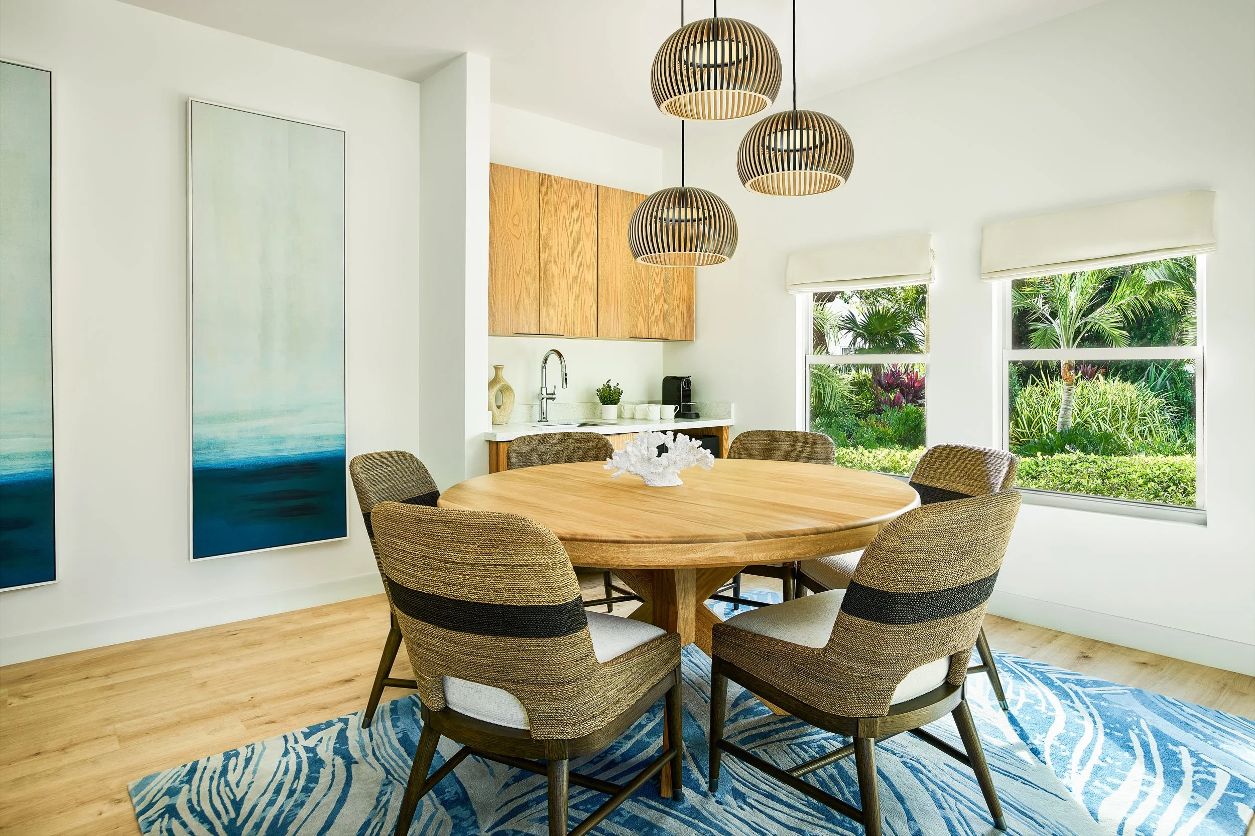 A modern dining room with a round wooden table surrounded by six upholstered chairs, a blue and white patterned rug, large windows with views of greenery, and a set of three wooden pendant lights hanging above the table.