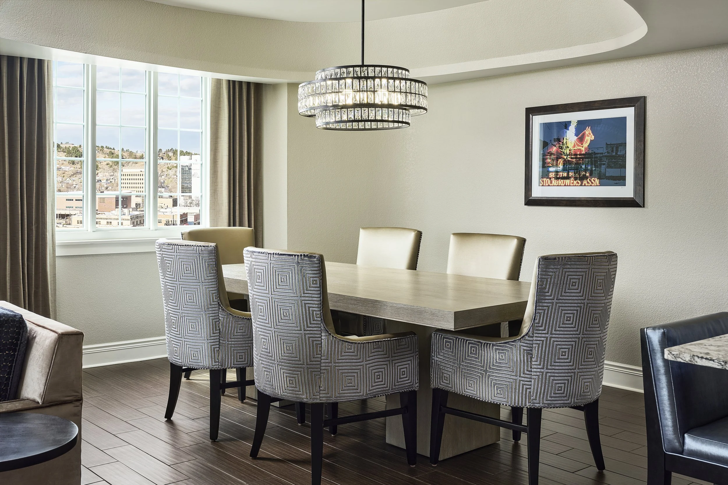 A dining room with a wooden table and chairs, a large window with curtains, and a chandelier hanging from the ceiling.