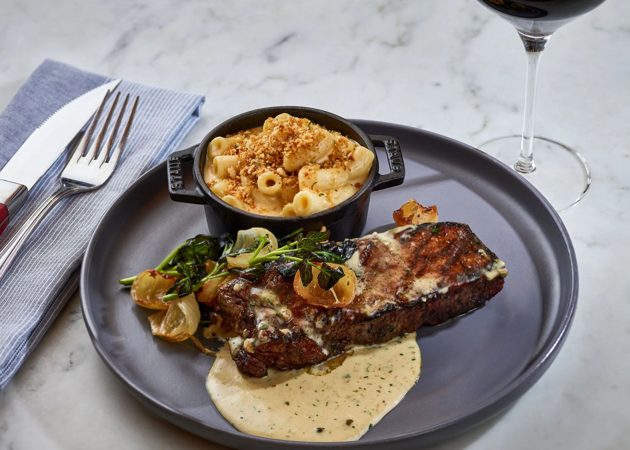A plate of food with mac and cheese topped with breadcrumbs, a side of greens, and a grilled meat with sauce, alongside a glass of red wine and silverware on a blue napkin.