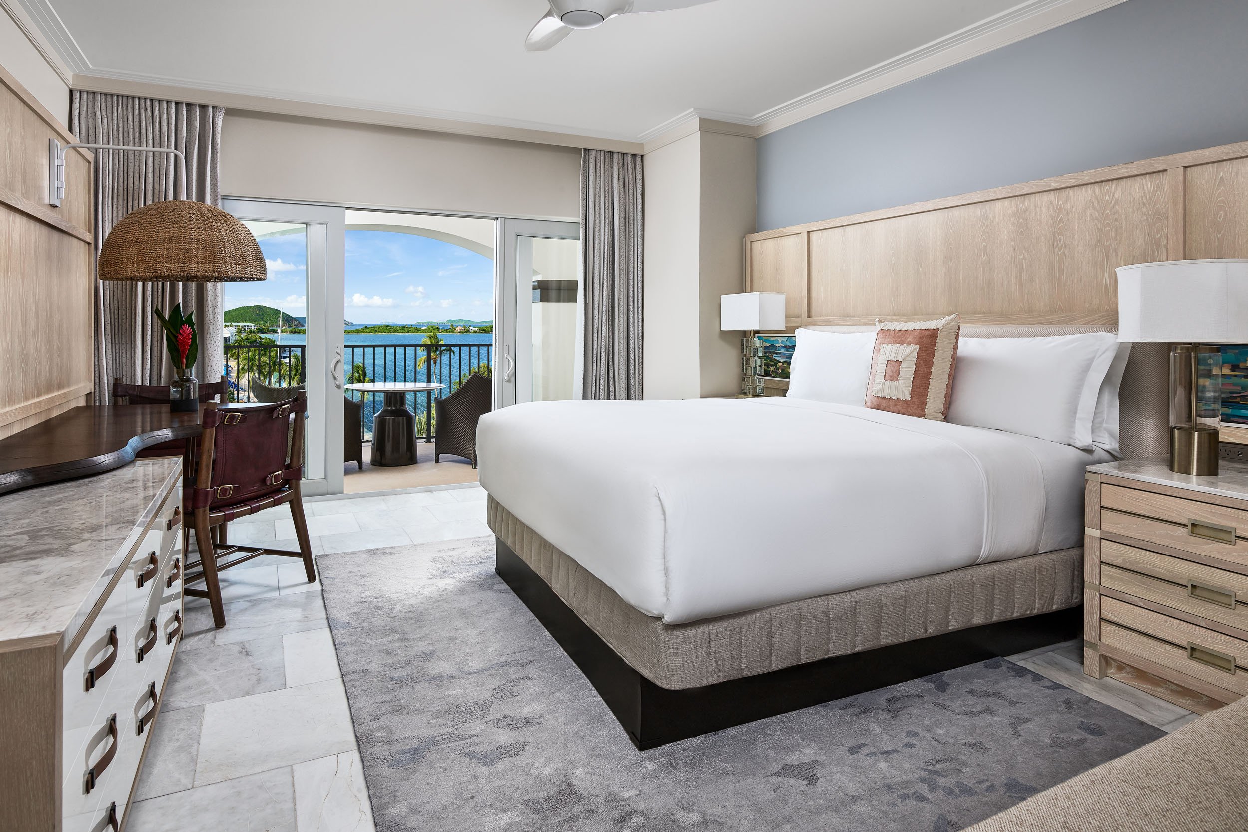 hotel room with a large bed, a view of water and greenery through a balcony door, a bedside lamp, a desk with a chair, and a ceiling fan.
