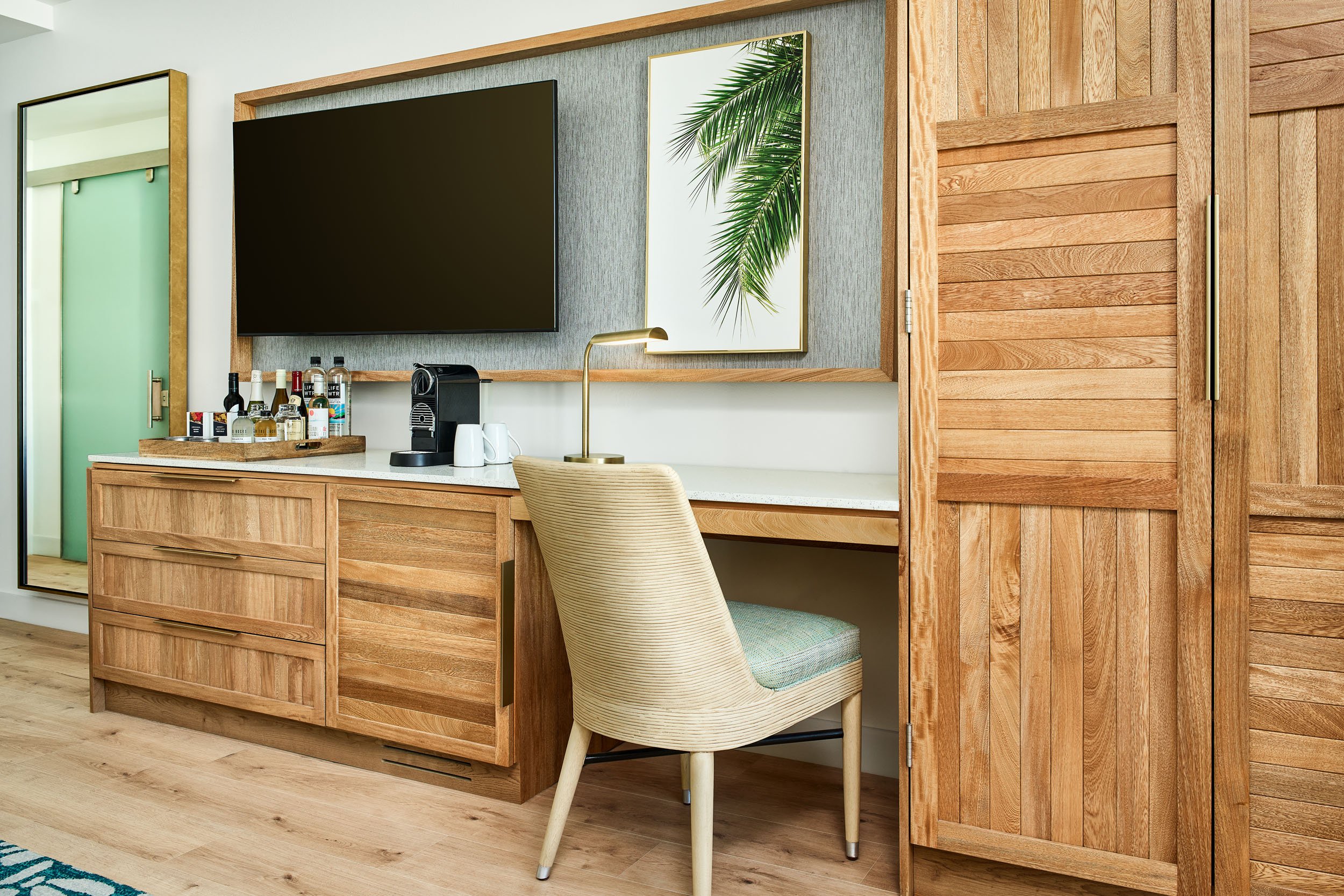 A coffee station in a room with wood accents, including a cabinet, mirror, and framed artwork of a palm tree, with a coffee machine, bottles, cups, and a lamp on the counter.