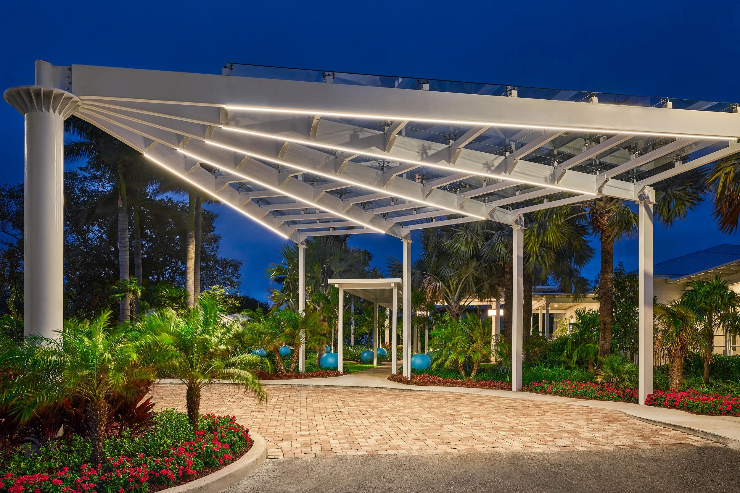 Night view of a modern outdoor structure with white beams and glass railings, illuminated by integrated lighting, surrounded by lush tropical plants and palm trees.