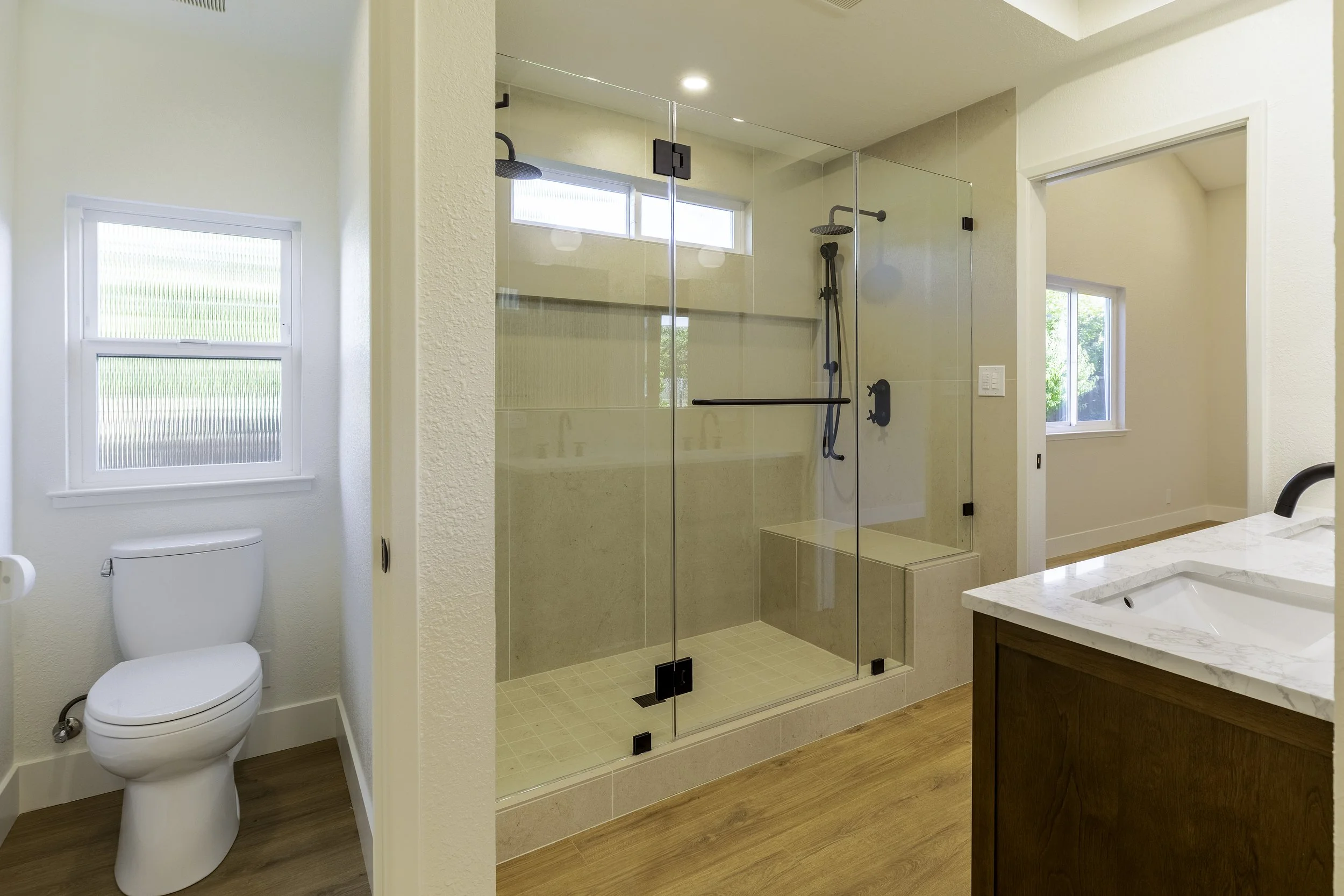 Modern bathroom with a glass shower enclosure, white walls, a window, and a wooden vanity with a marble top.