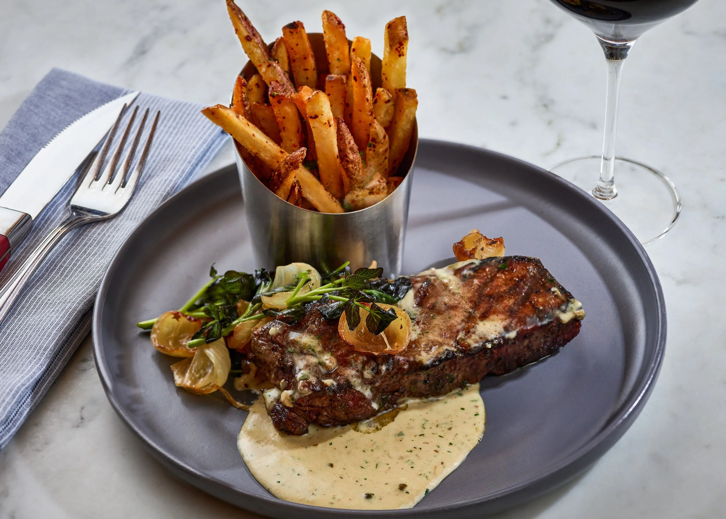 A plate with grilled steak topped with sauce and vegetables, a metal container of French fries, and a glass of red wine on a marble table.