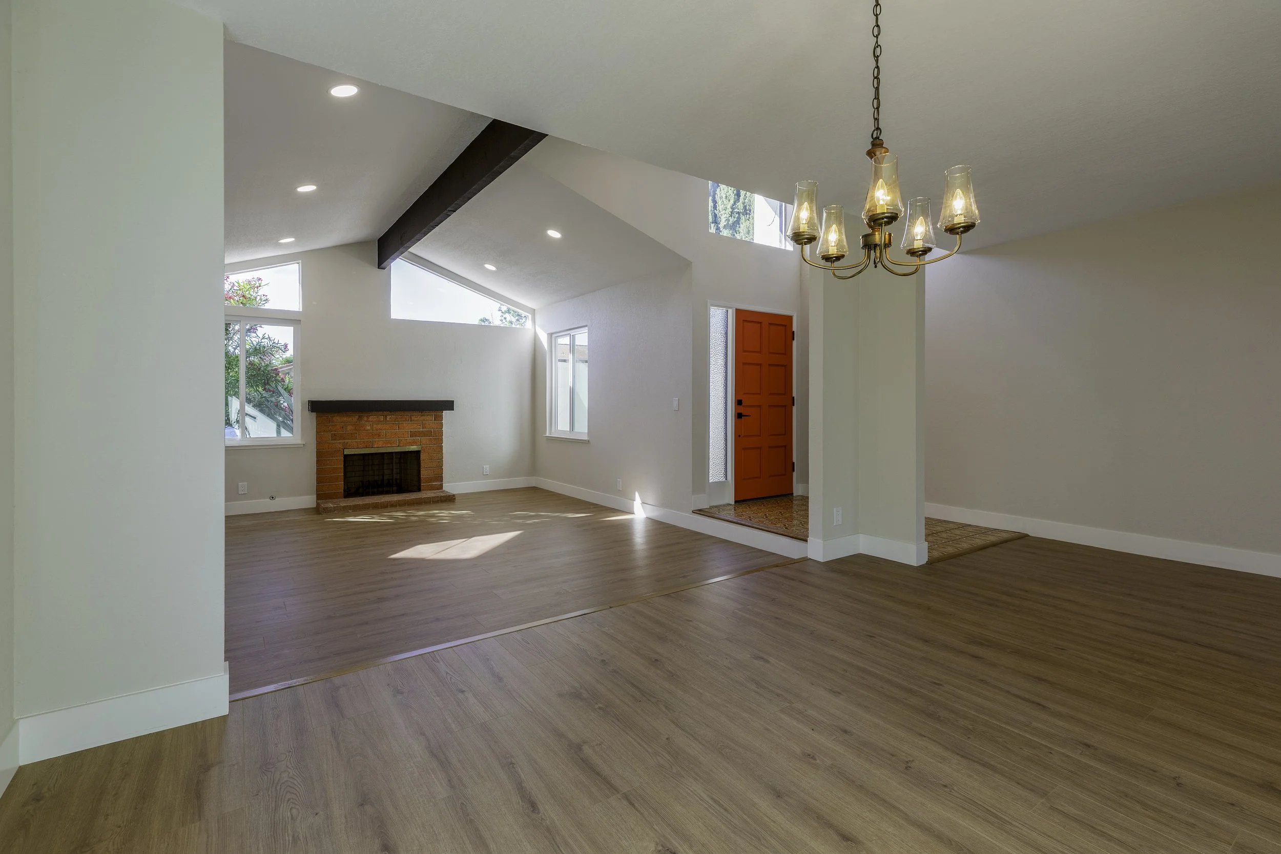Empty living room with hardwood floors, white walls, a brick fireplace, multiple windows, a red door, and a chandelier.