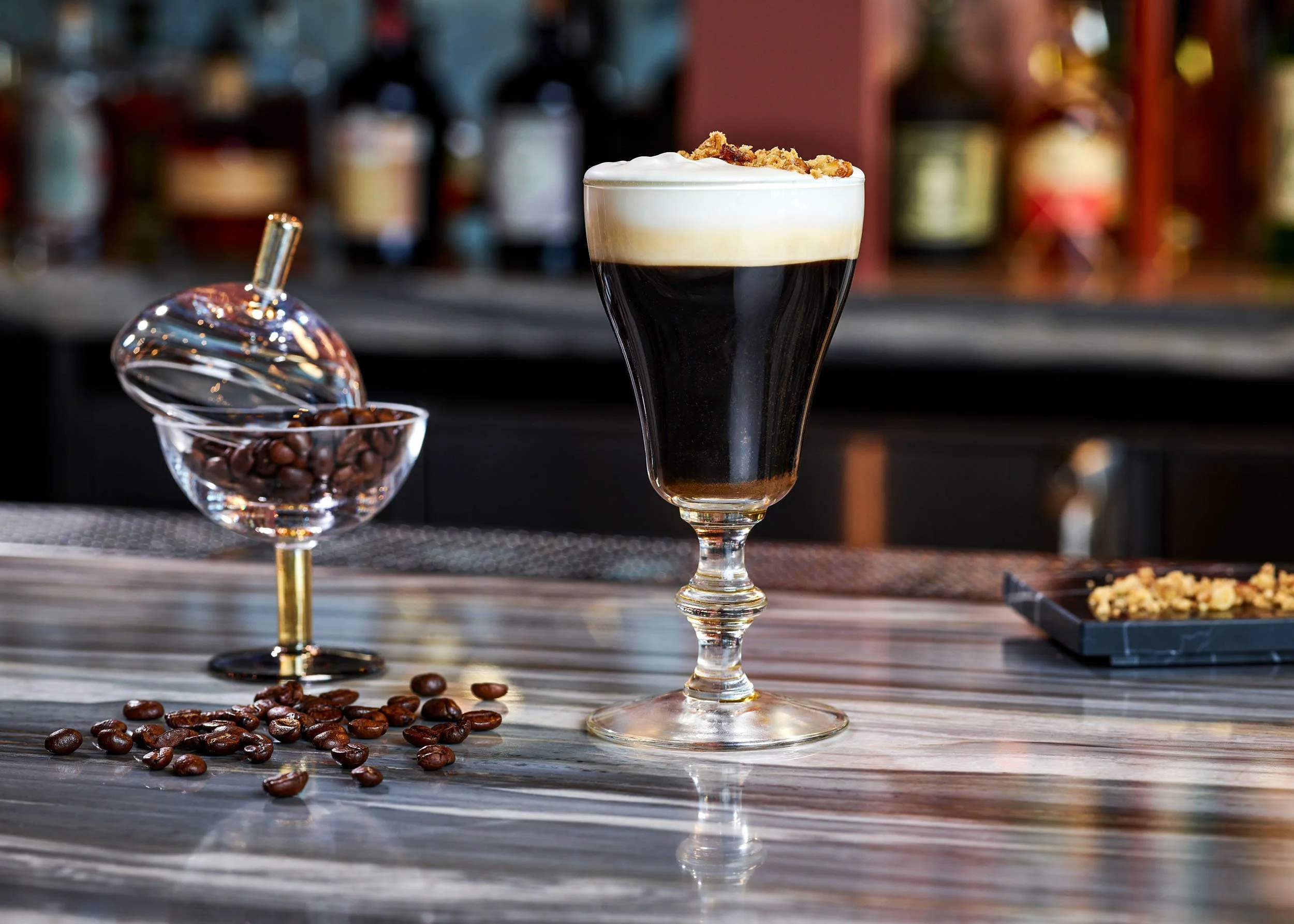 A coffee cocktail garnished with whipped cream and crushed graham crackers, served in a tall glass, with coffee beans and an espresso spoon beside it on a wooden surface.