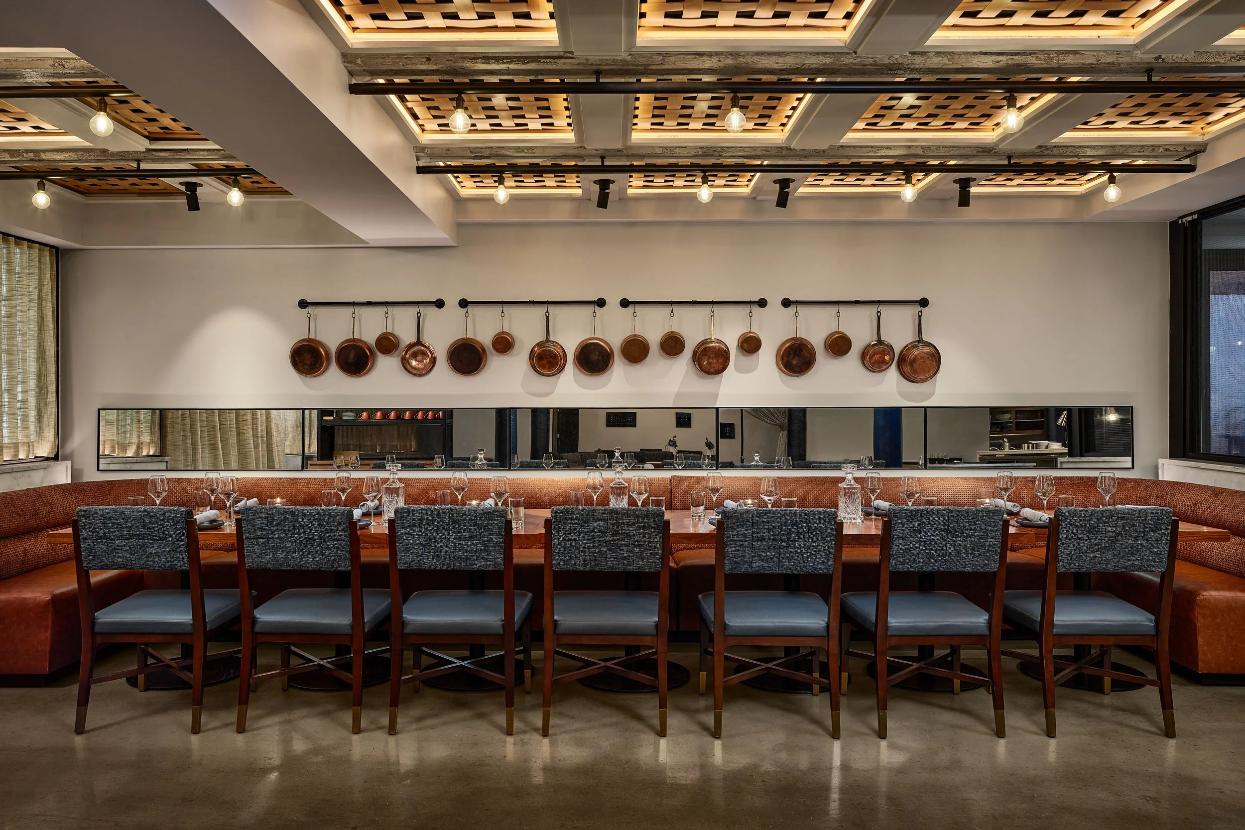 A modern restaurant interior with a long dining table set with wine glasses and napkins, a sofa with chairs along it, and copper pots hanging on a white wall. The ceiling has wooden panels and hanging light bulbs.