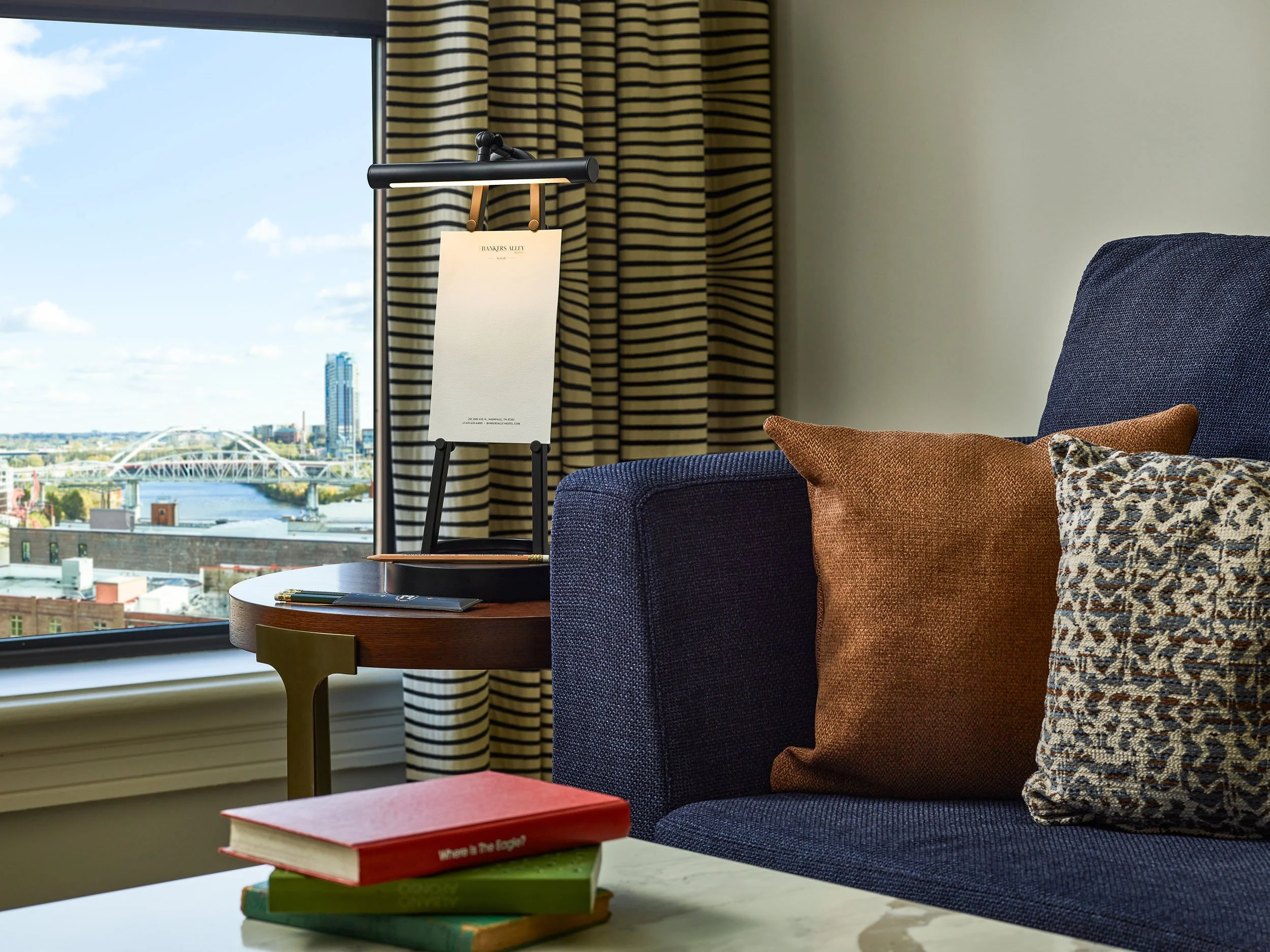 A hotel room with a large window showing a cityscape with a bridge over a river. Inside, there are striped curtains, a dark blue armchair with brown and leopard print pillows, a small round table with a black tray and a notebook, and a stack of books