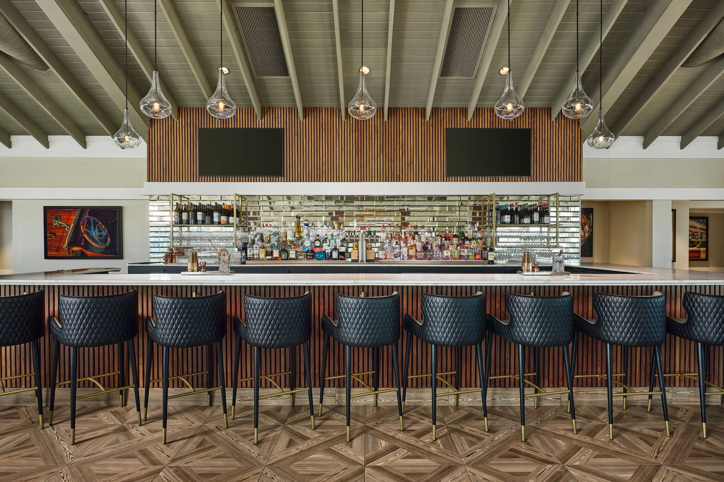 Modern bar with a wooden and mirrored back wall, black bar stools, and glass pendant lights hanging from a vaulted ceiling.