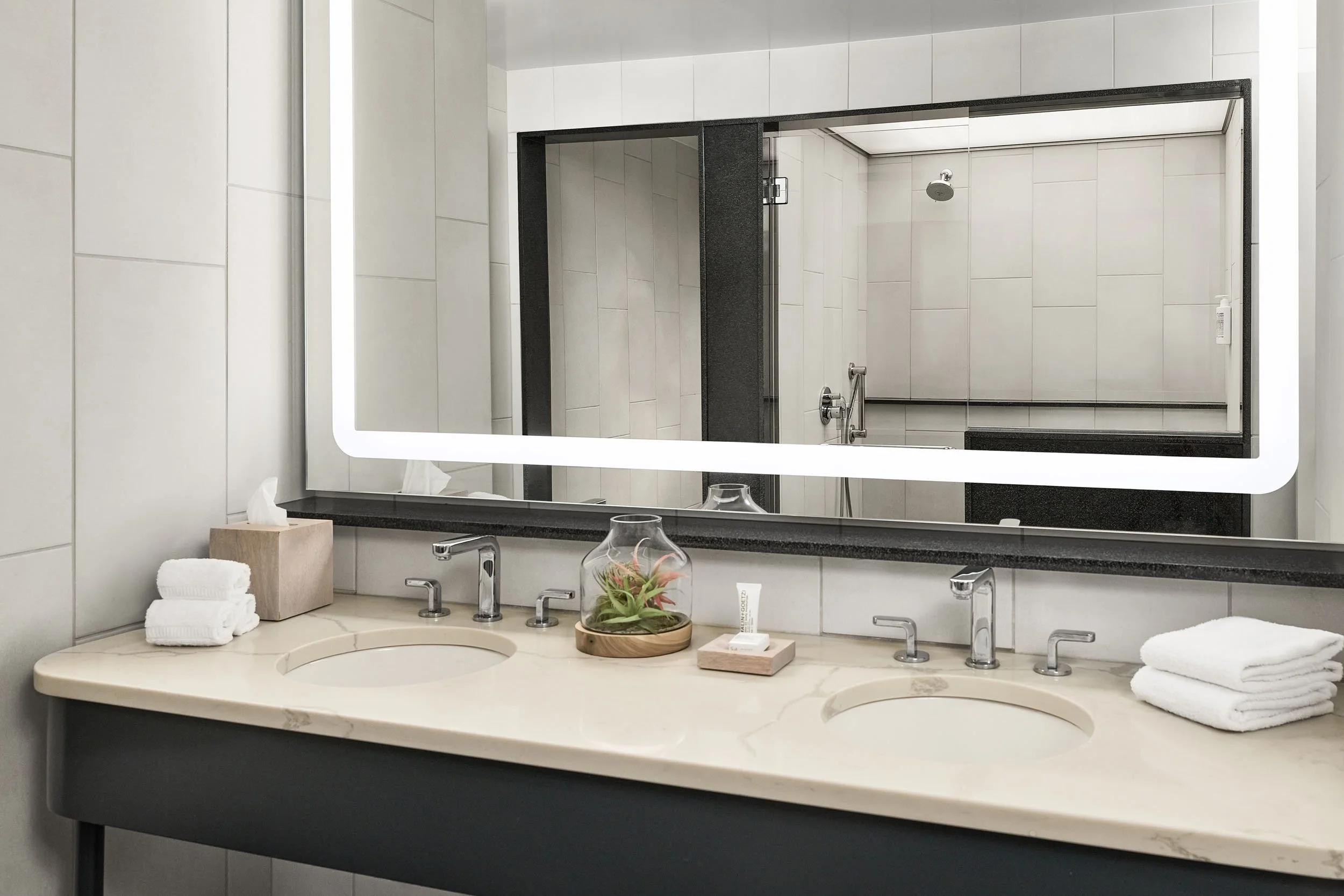 Two white bathroom sinks with a marble countertop, each with a silver faucet. On the left side, there are folded white towels and a beige tissue box. In the center, a small potted plant and a tube of skincare products are placed. Behind the mirror, a