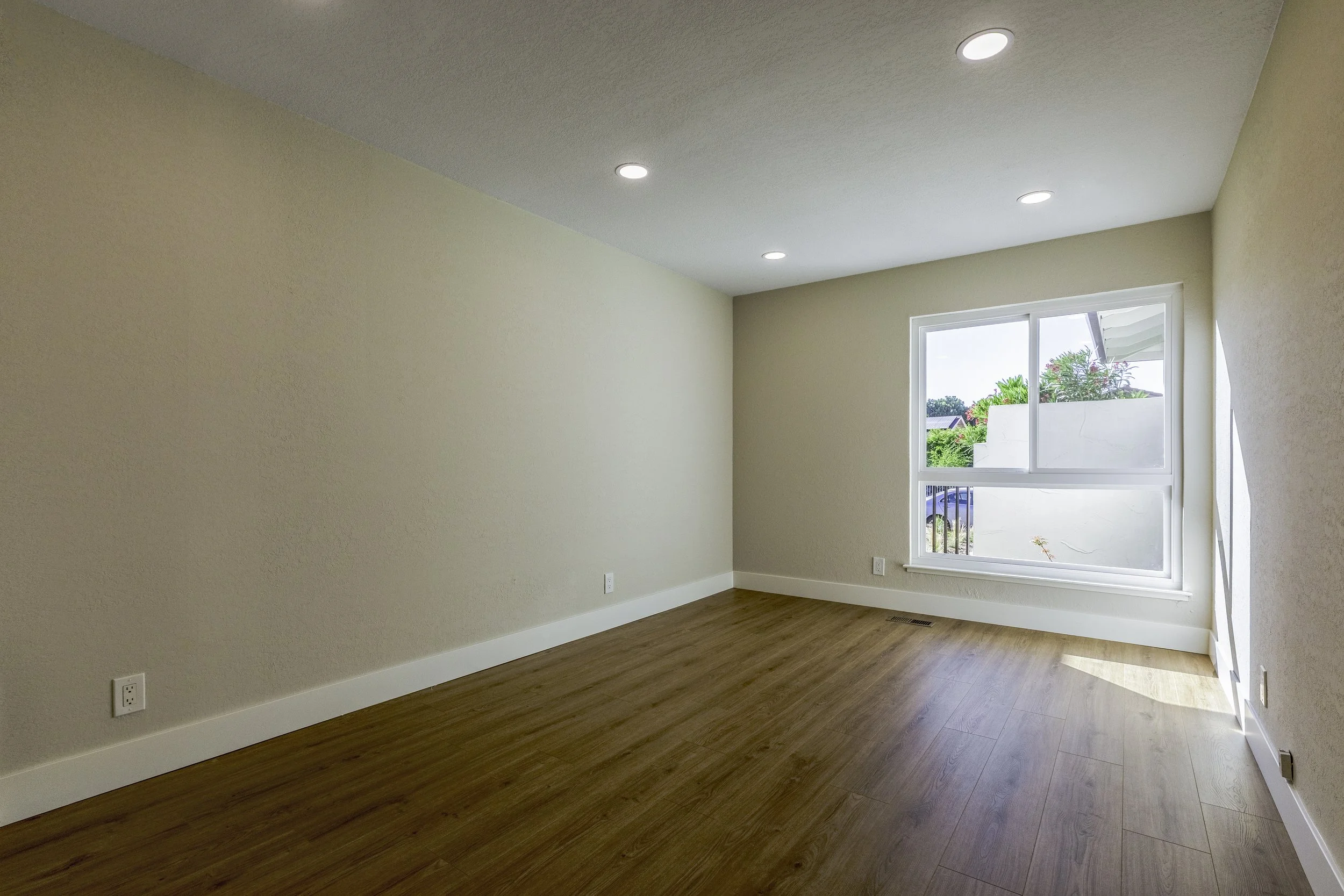 Empty room with beige walls, wooden flooring, a large window showing outdoor greenery, and ceiling lights.
